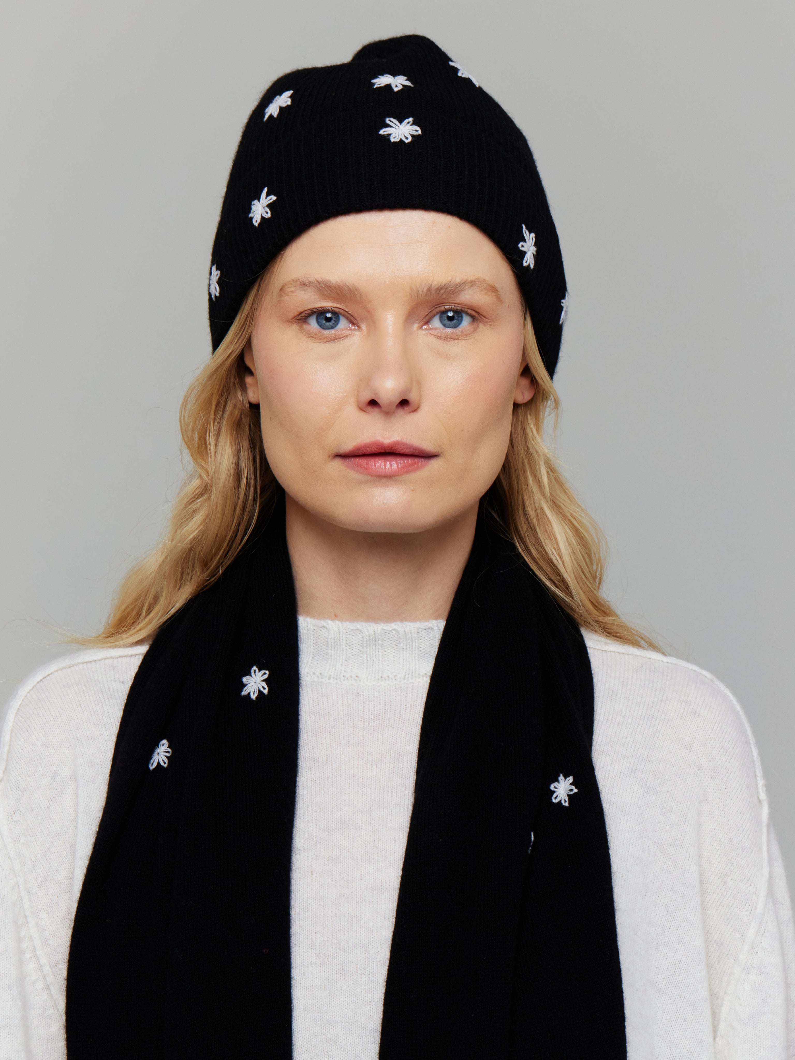 A woman wearing a black cashmere hat with white embroidered flowers on it, paired with a white sweater and matching scarf.