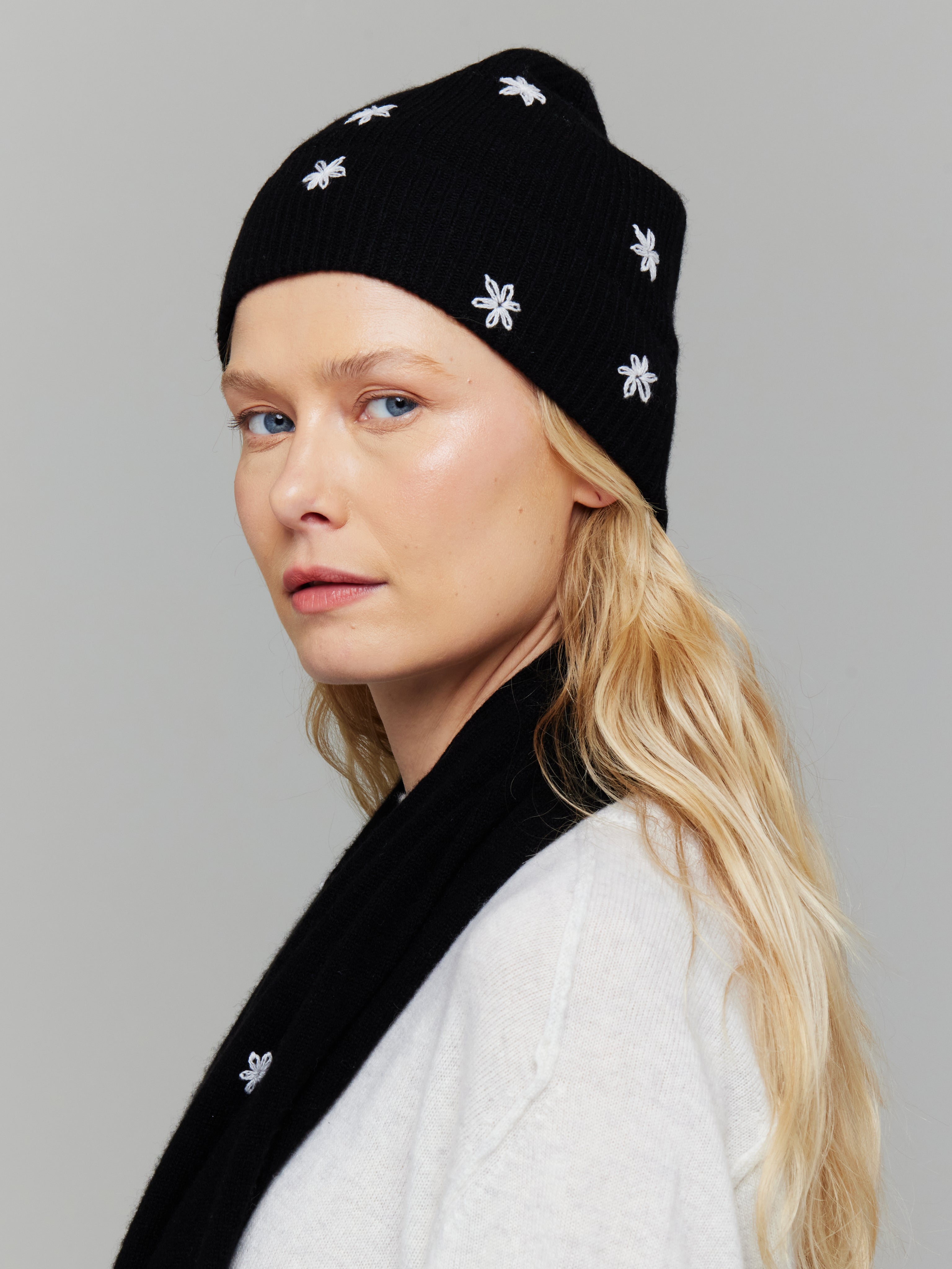 A woman wearing a black cashmere hat with white embroidered flowers on it, paired with a white sweater and matching scarf.