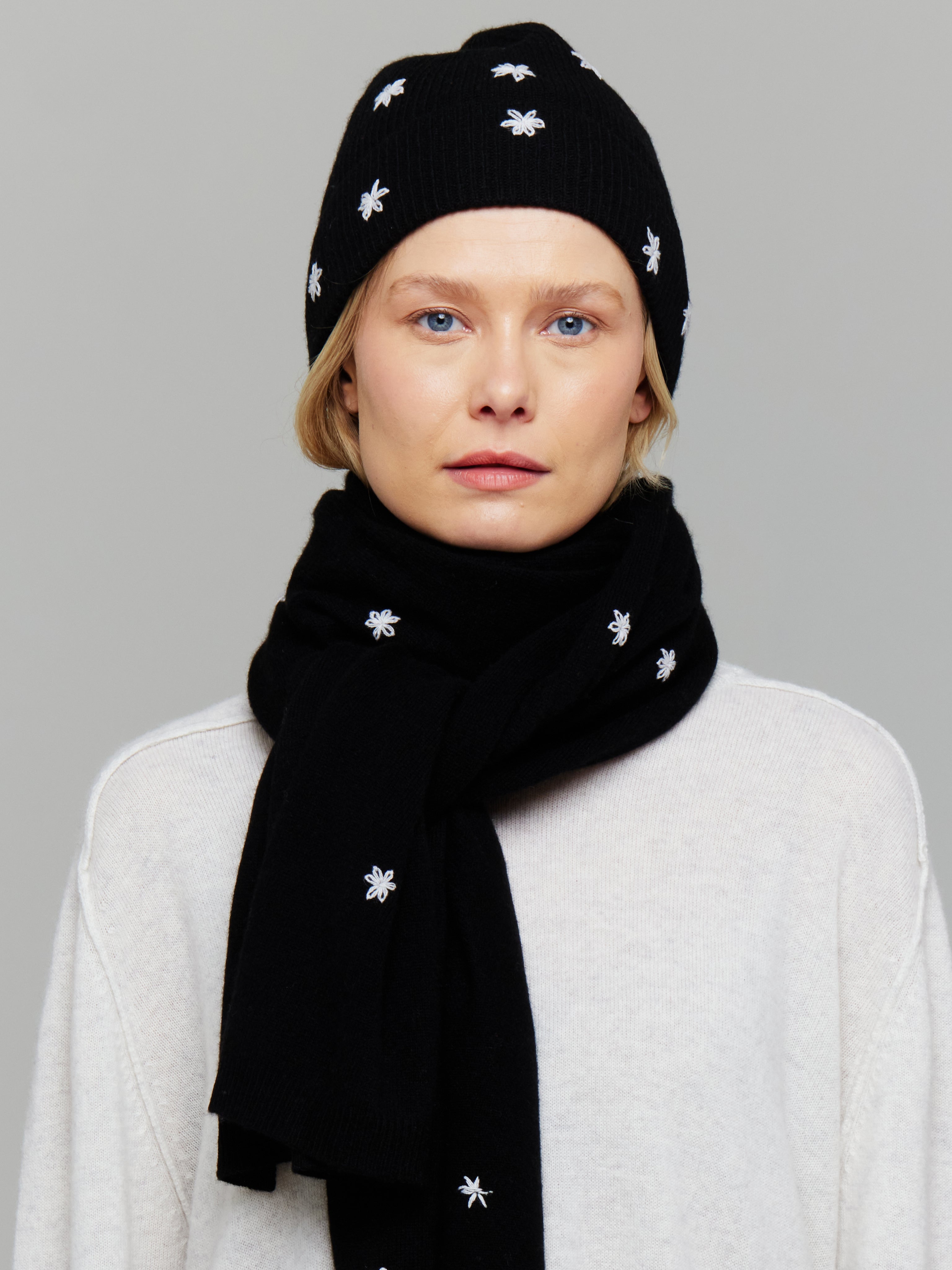 A woman wearing a black cashmere hat with white embroidered flowers on it, paired with a white sweater and matching scarf.