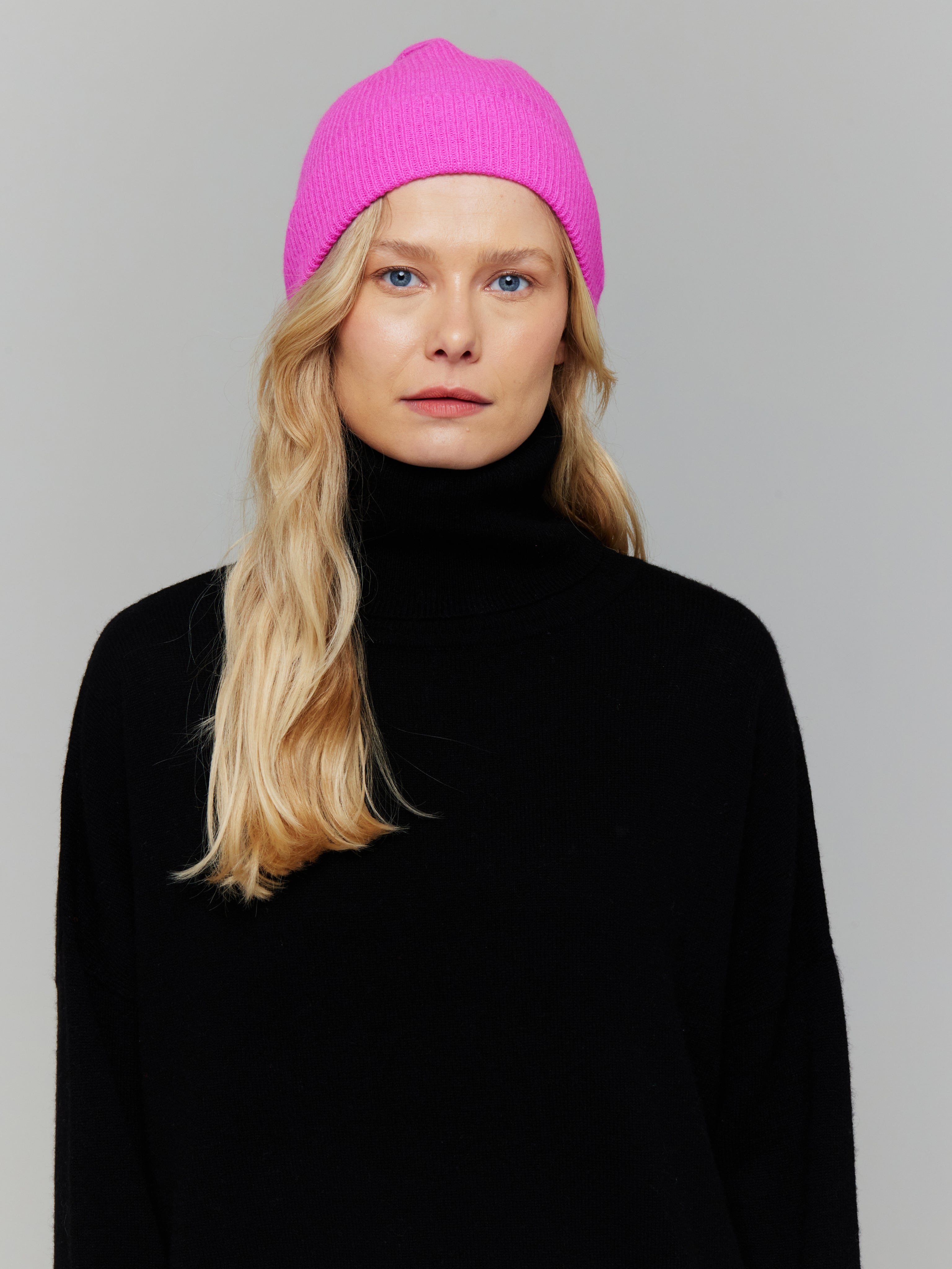 Person wearing a bright pink beanie and black turtleneck against a gray background