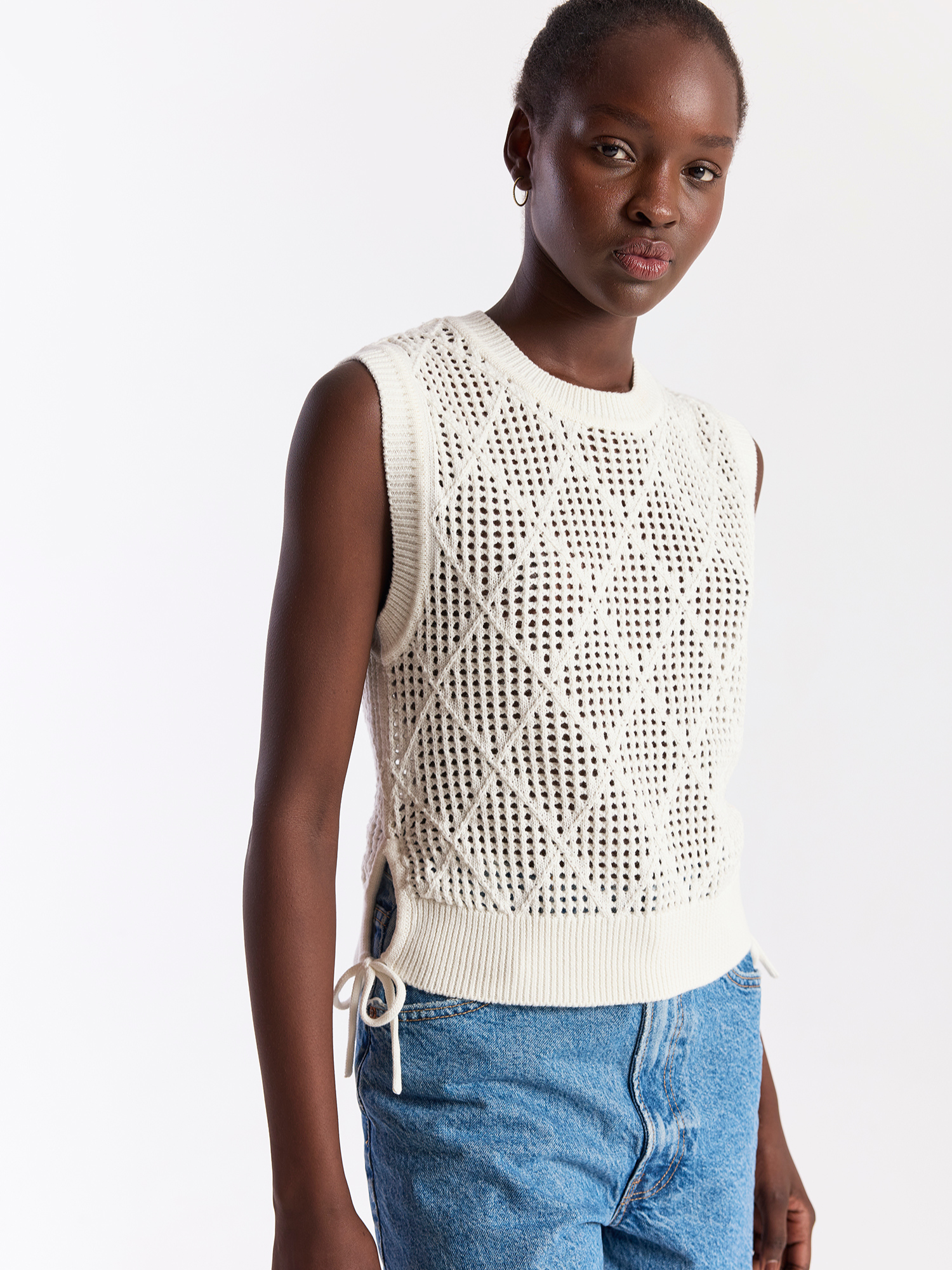 Person wearing a white sleeveless knitted top and blue jeans on a light gray background