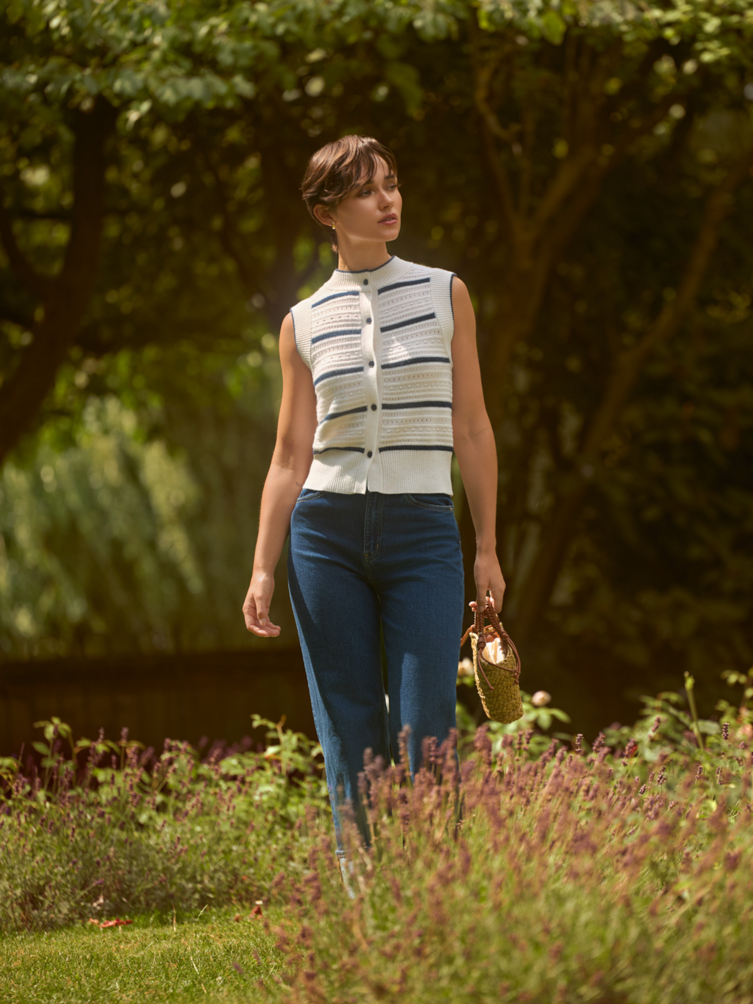 Woman in a striped sleeveless top and blue jeans standing in a garden.