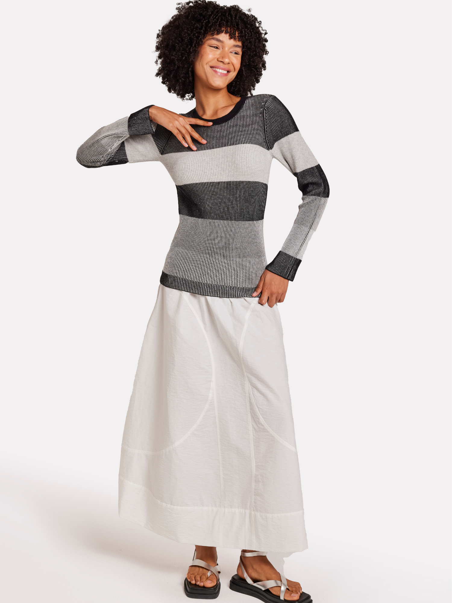 A model wearing a grey striped crew neck sweater with long sleeves, featuring black, white and grey stripes with a white skirt.