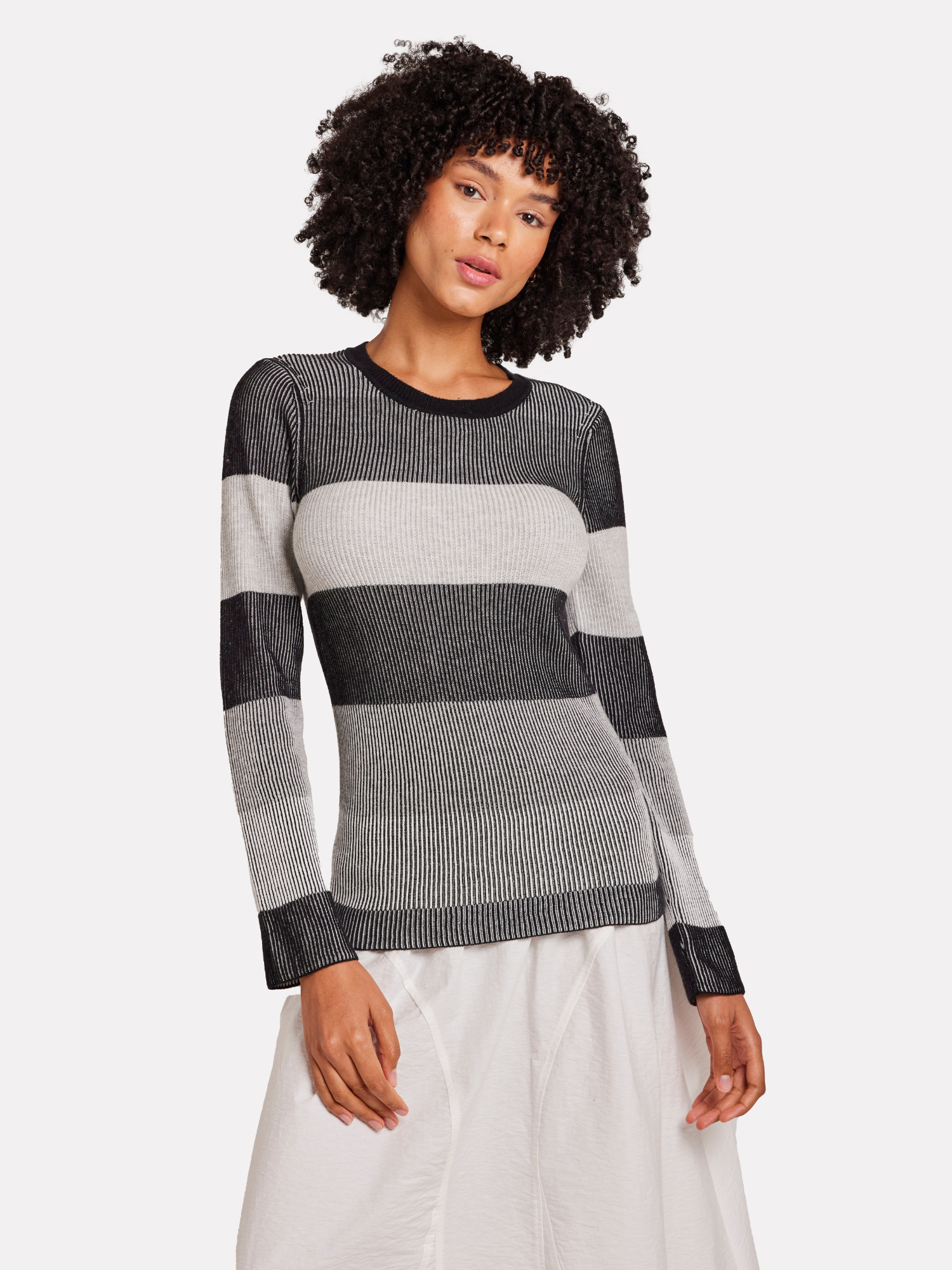 A model wearing a grey striped crew neck sweater with long sleeves, featuring black, white and grey stripes with a white skirt.
