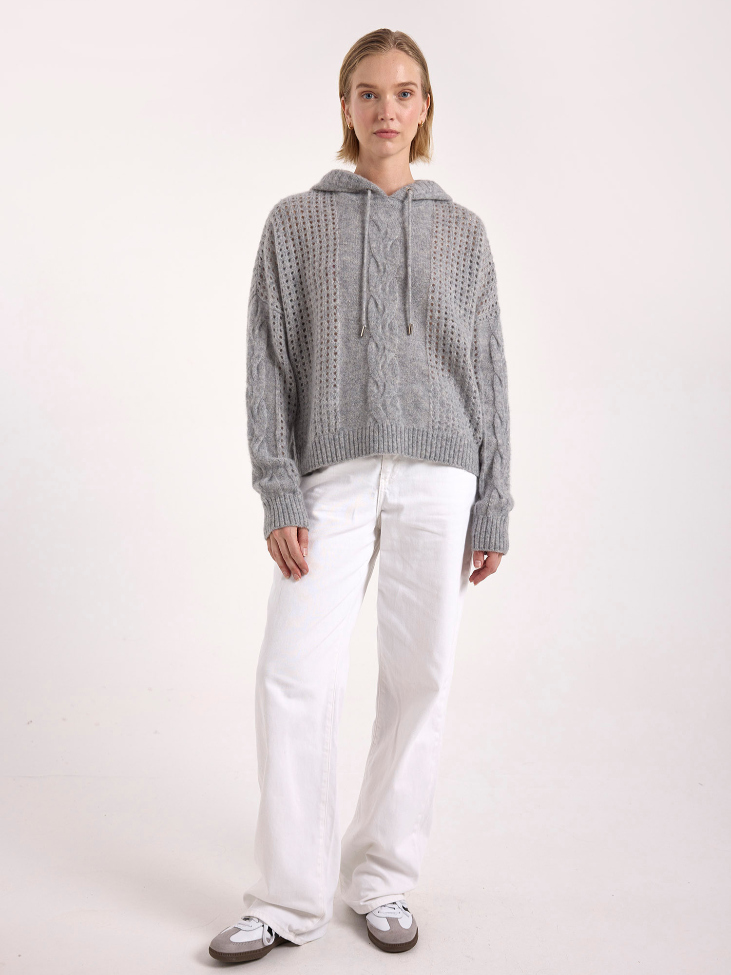 Woman wearing a grey cable knit cashmere sweater and white pants on a white background