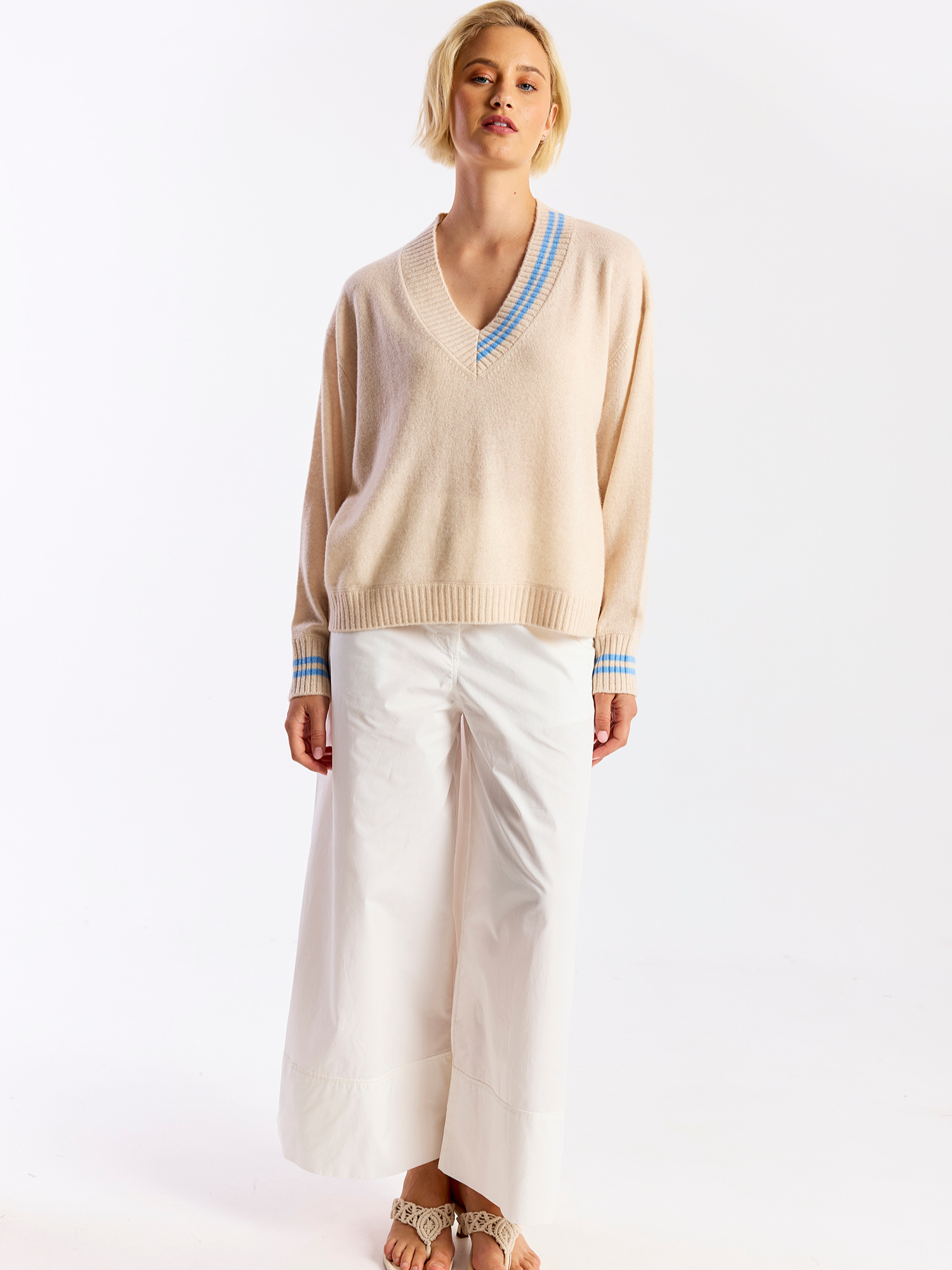 Woman wearing a beige sweater with blue trim and white pants on a white background