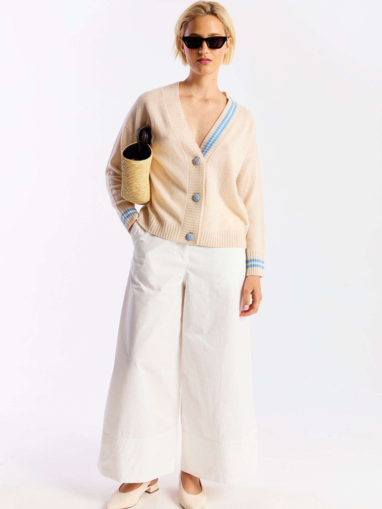 Woman wearing a beige cardigan with blue trim, white pants, and holding a woven bag on a white background