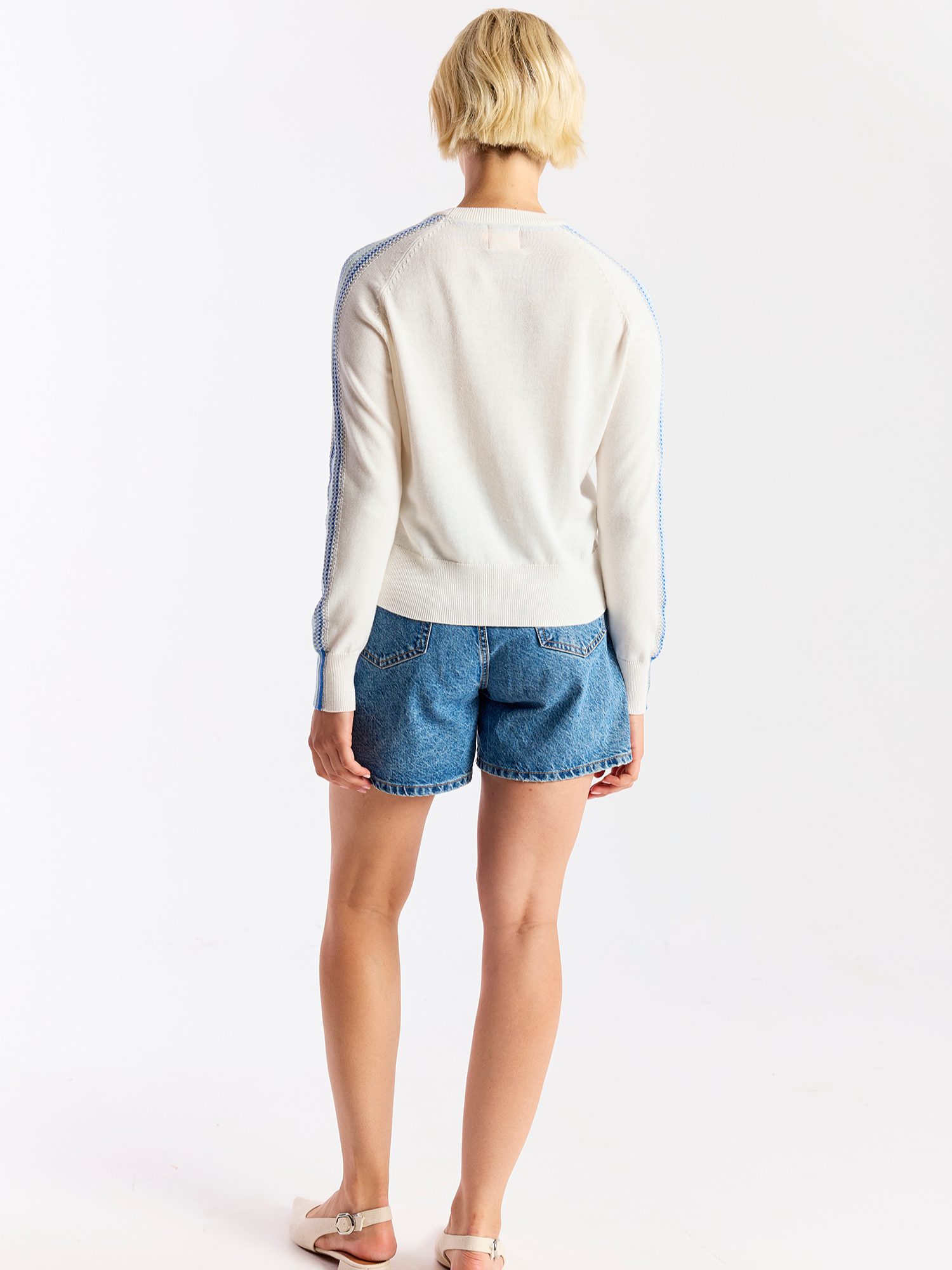 Person wearing a white sweater with blue accents and denim shorts on a white background