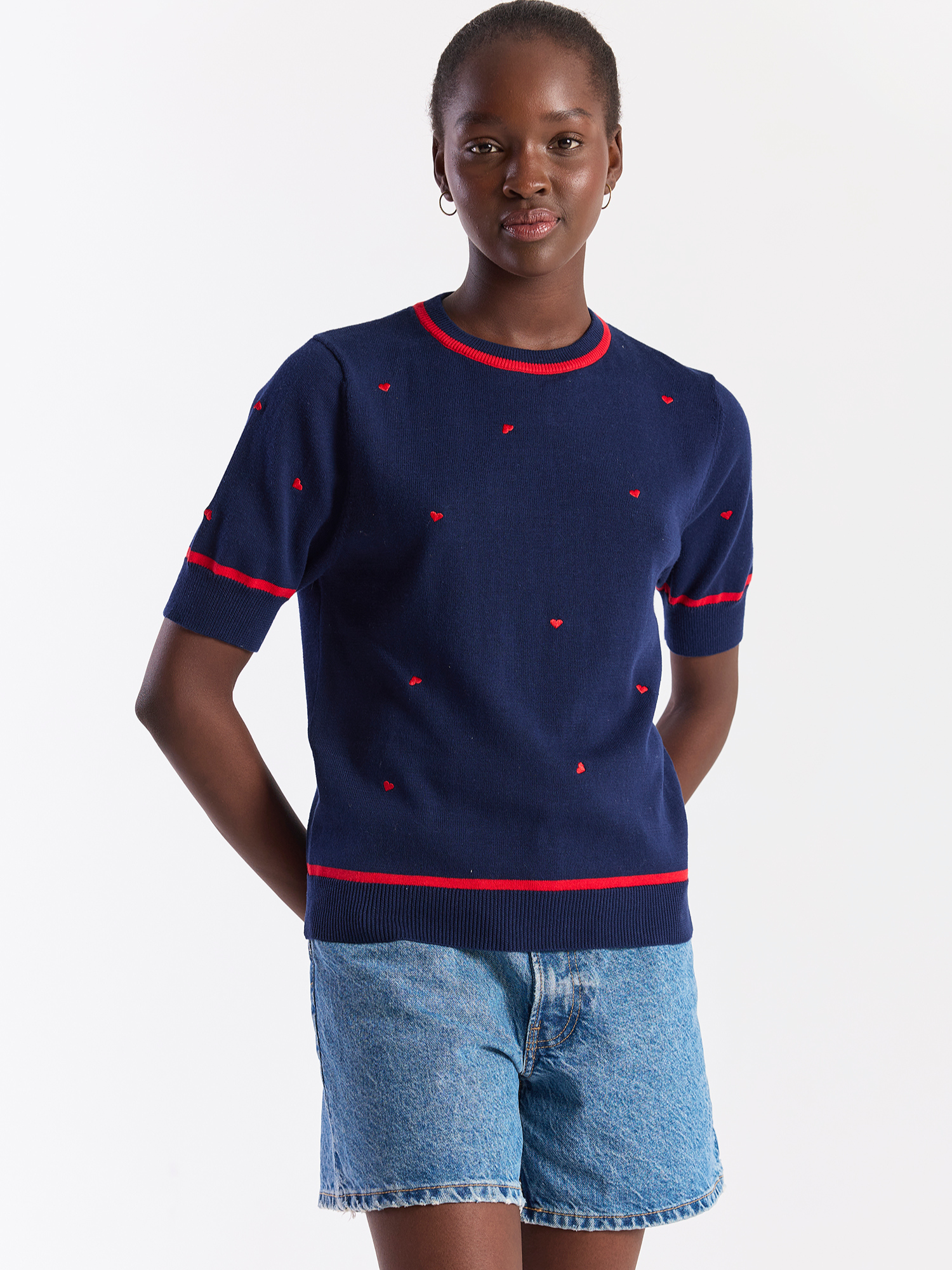 Person wearing a navy sweater with red accents and denim shorts on a white background