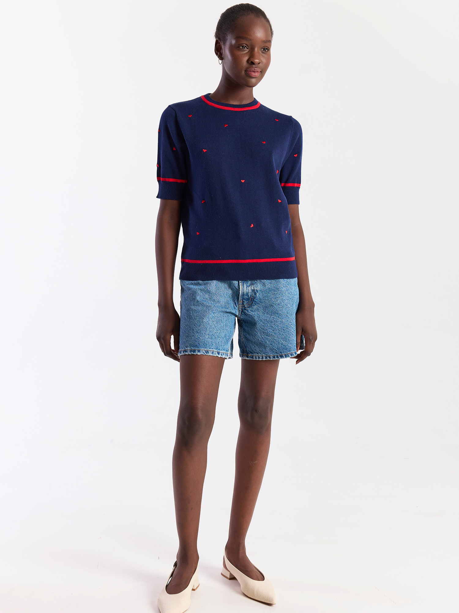 Woman wearing a navy sweater with red trim and denim shorts on a white background