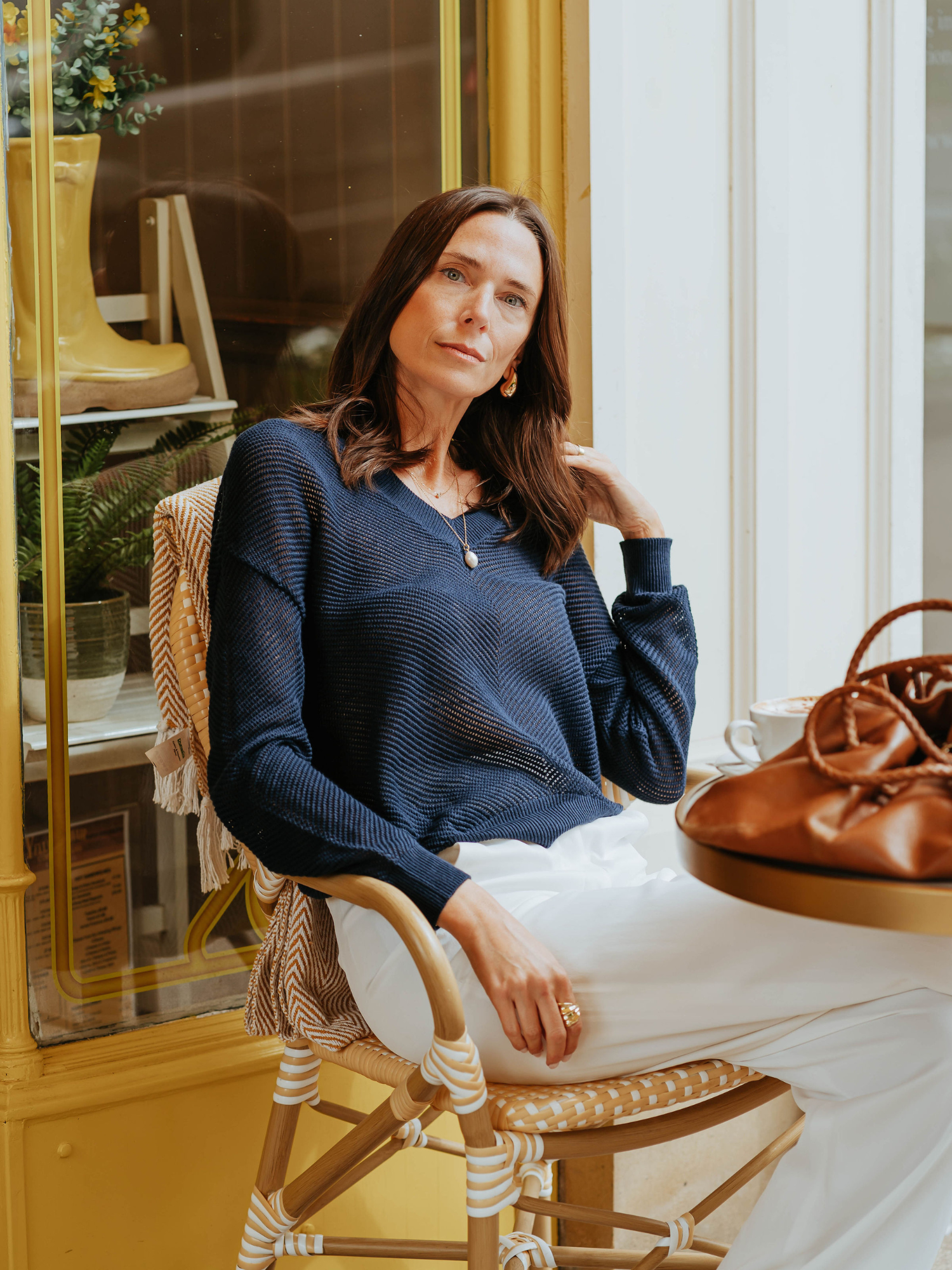 A woman wearing a navy V-neck jumper with pointelle chevron detail with white trousers sat outside a cafe
