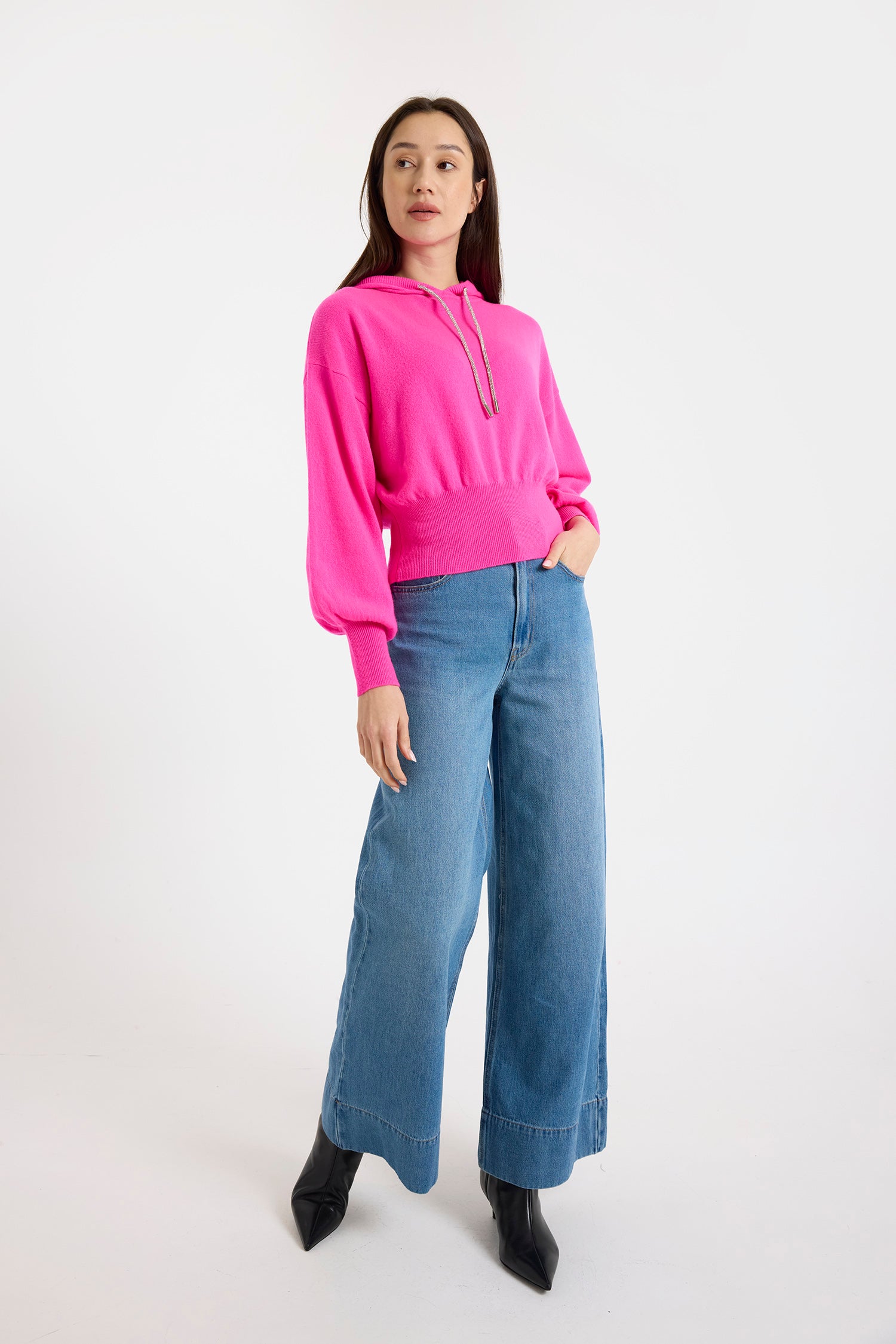 A woman wearing a pink cashmere hoodie with rhinestone drawstrings and blue jeans over a white background.