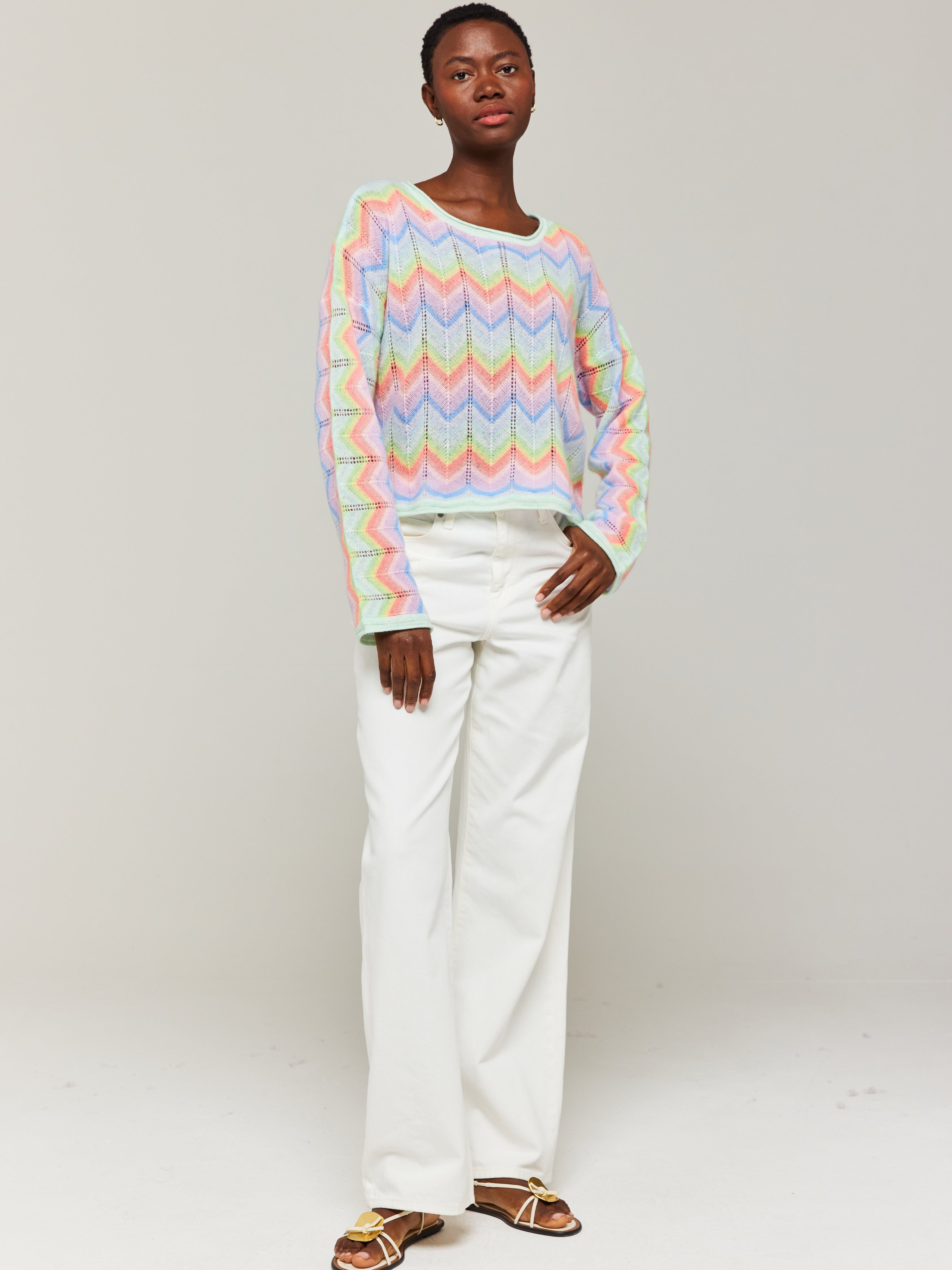 A person wearing an oversized multicolored cashmere sweater with a slash neck, paired with white trousers.