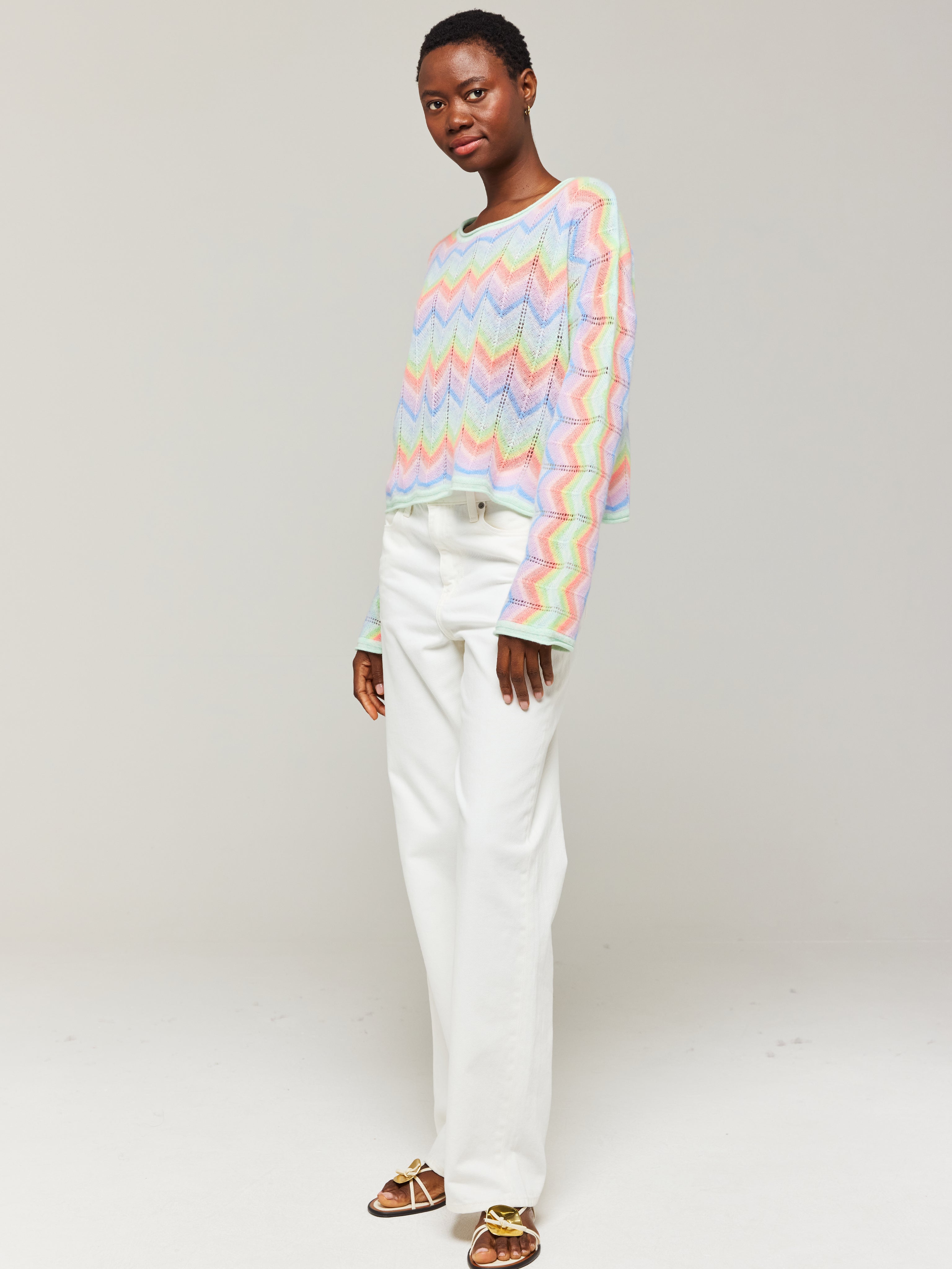 A person wearing an oversized multicolored cashmere sweater with a slash neck, paired with white trousers.