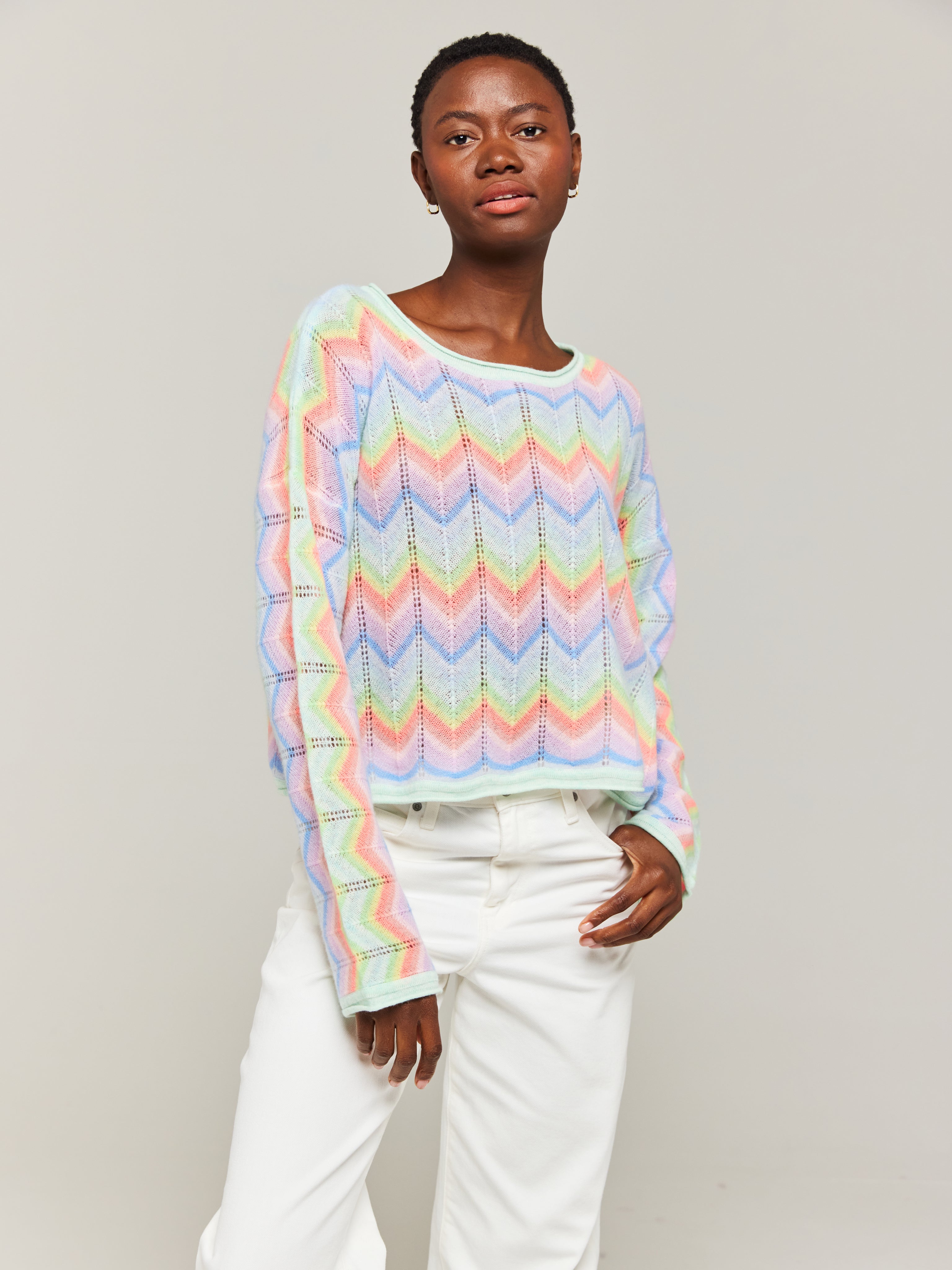 A person wearing an oversized multicolored cashmere sweater with a slash neck, paired with white trousers.