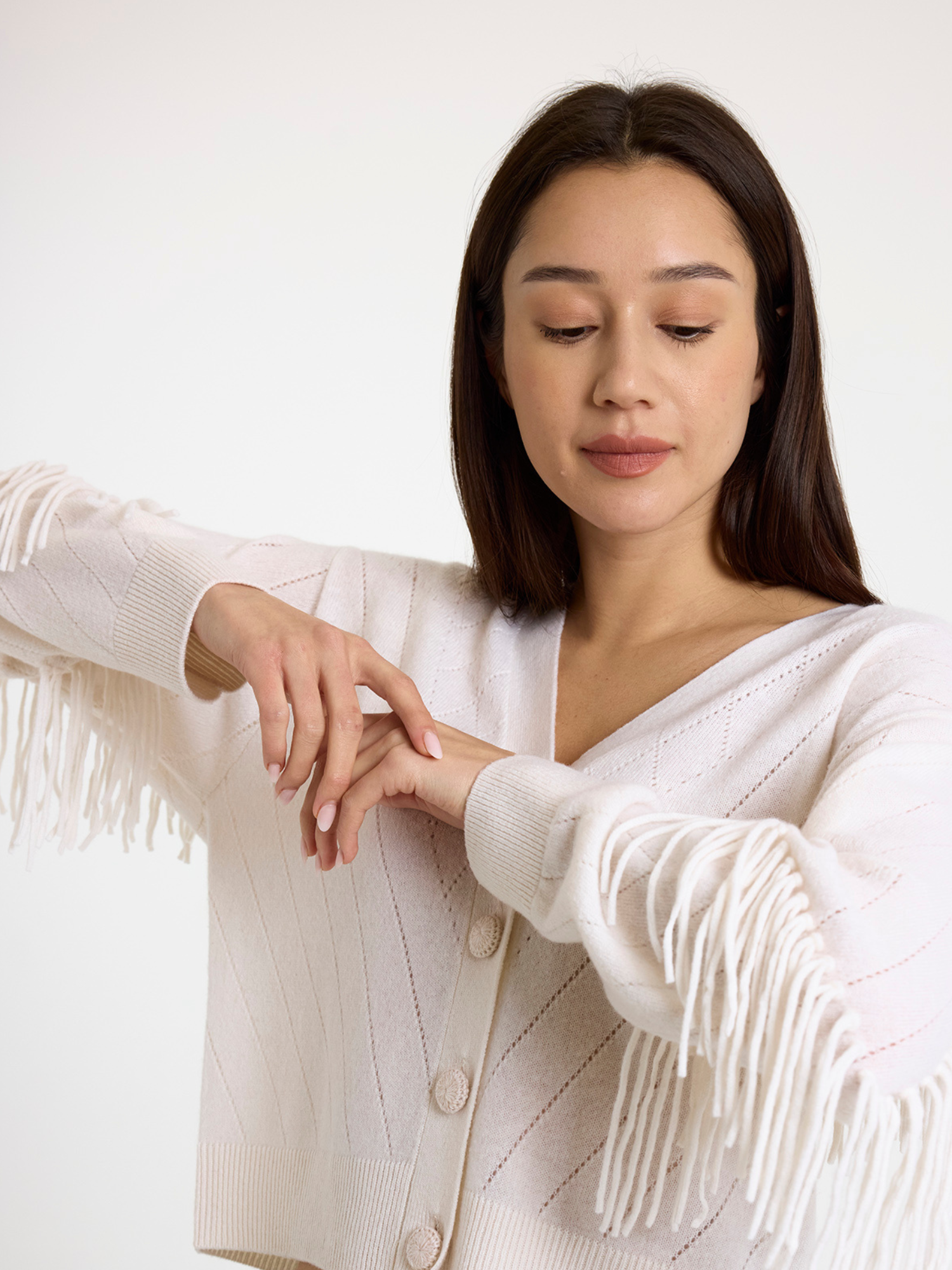 Woman wearing a white cardigan with fringe details on a plain background