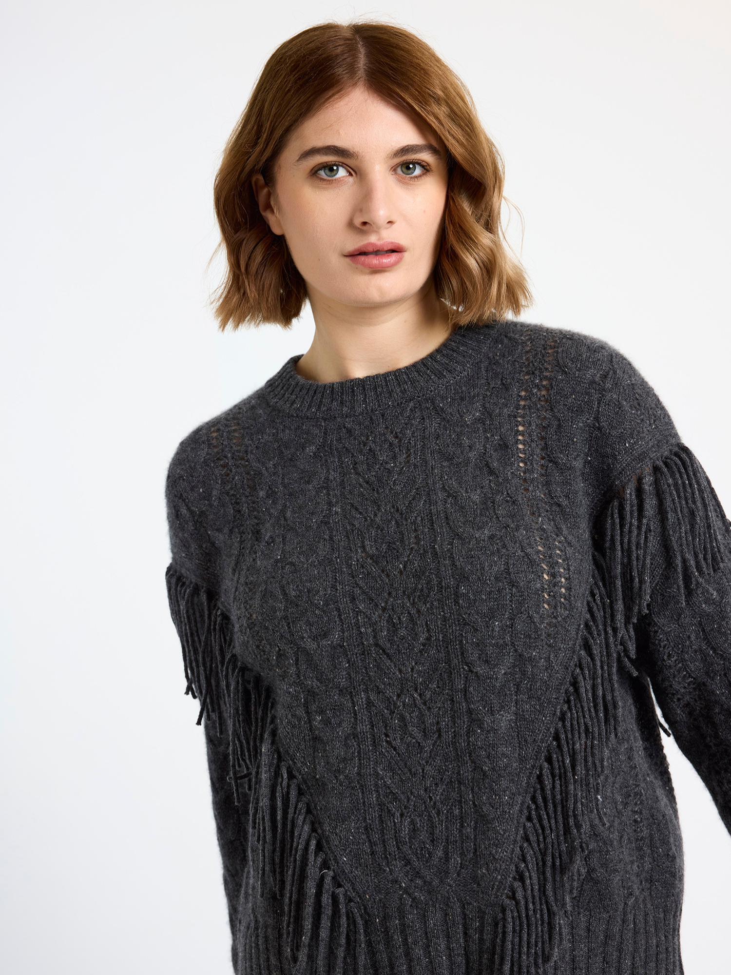 Woman wearing a dark grey cable sweater with fringe details on a light background