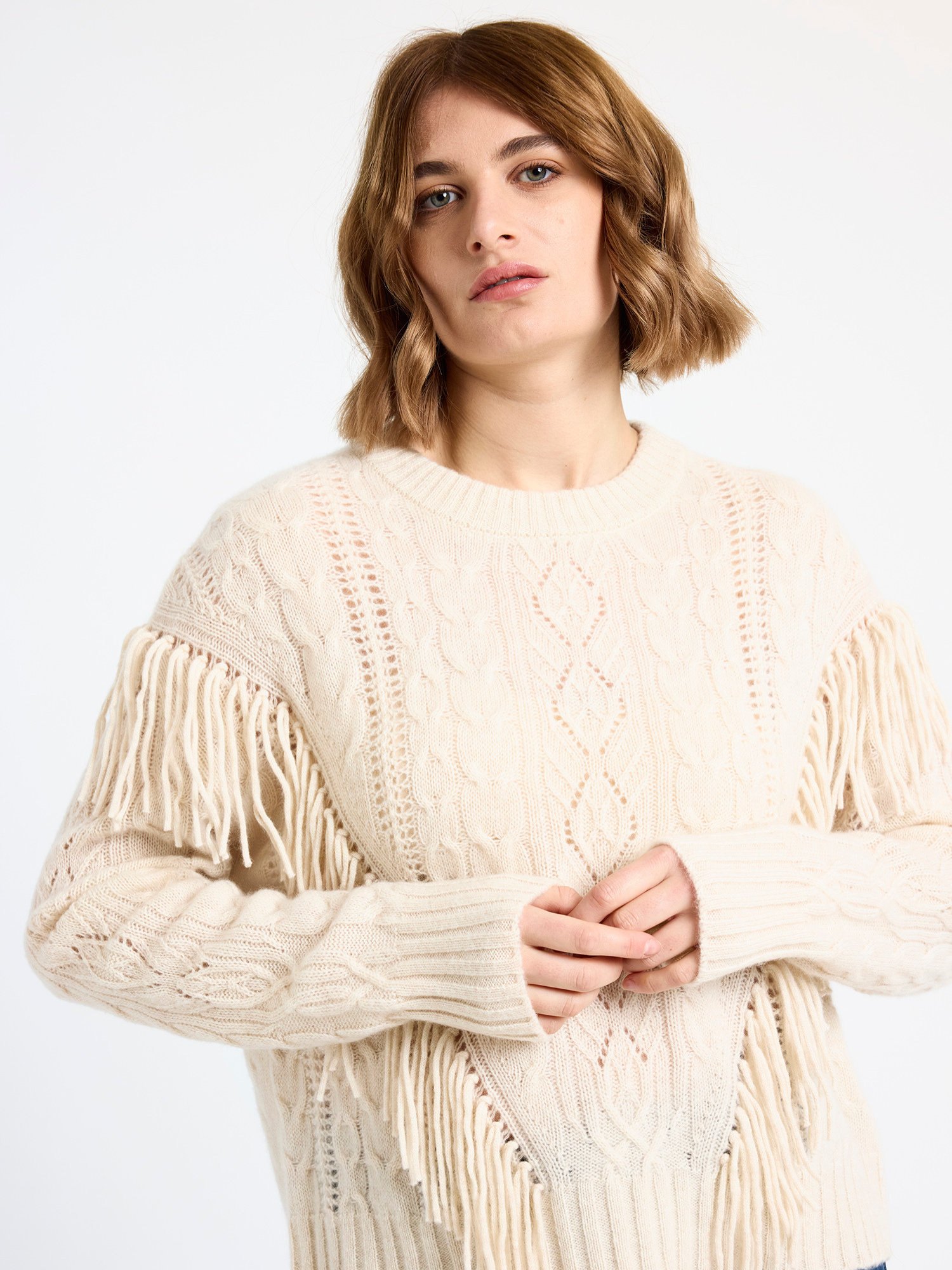 Woman wearing a cream-colored cable sweater with fringe details on a white background