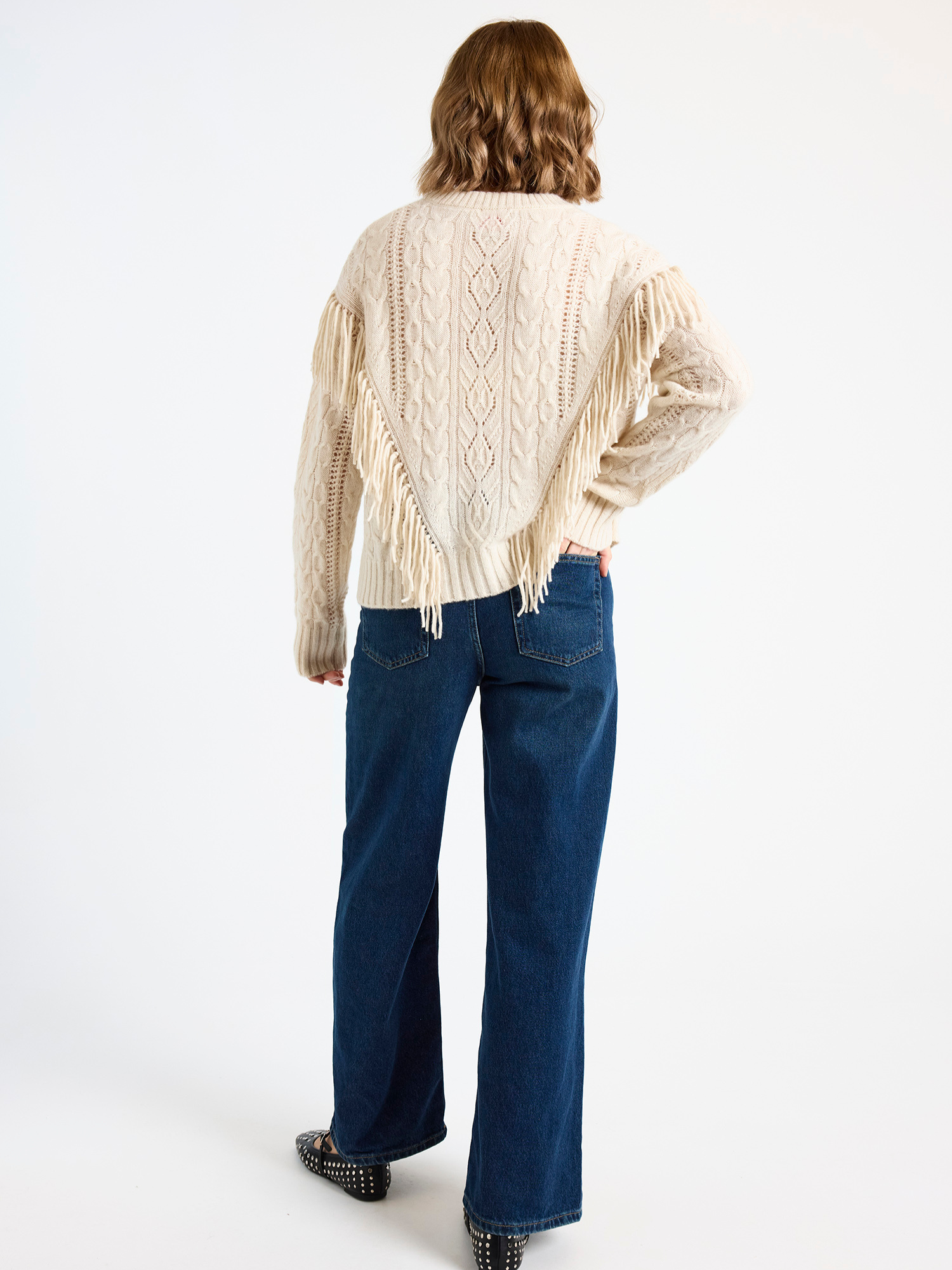 Woman wearing a cream cable knit sweater with fringe detail and blue jeans on a white background