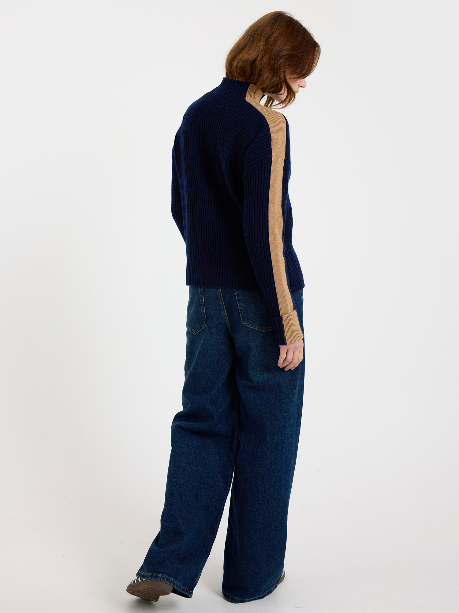 Woman wearing a navy ribbed sweater with camel colour stripe down the arm and blue jeans on a white background