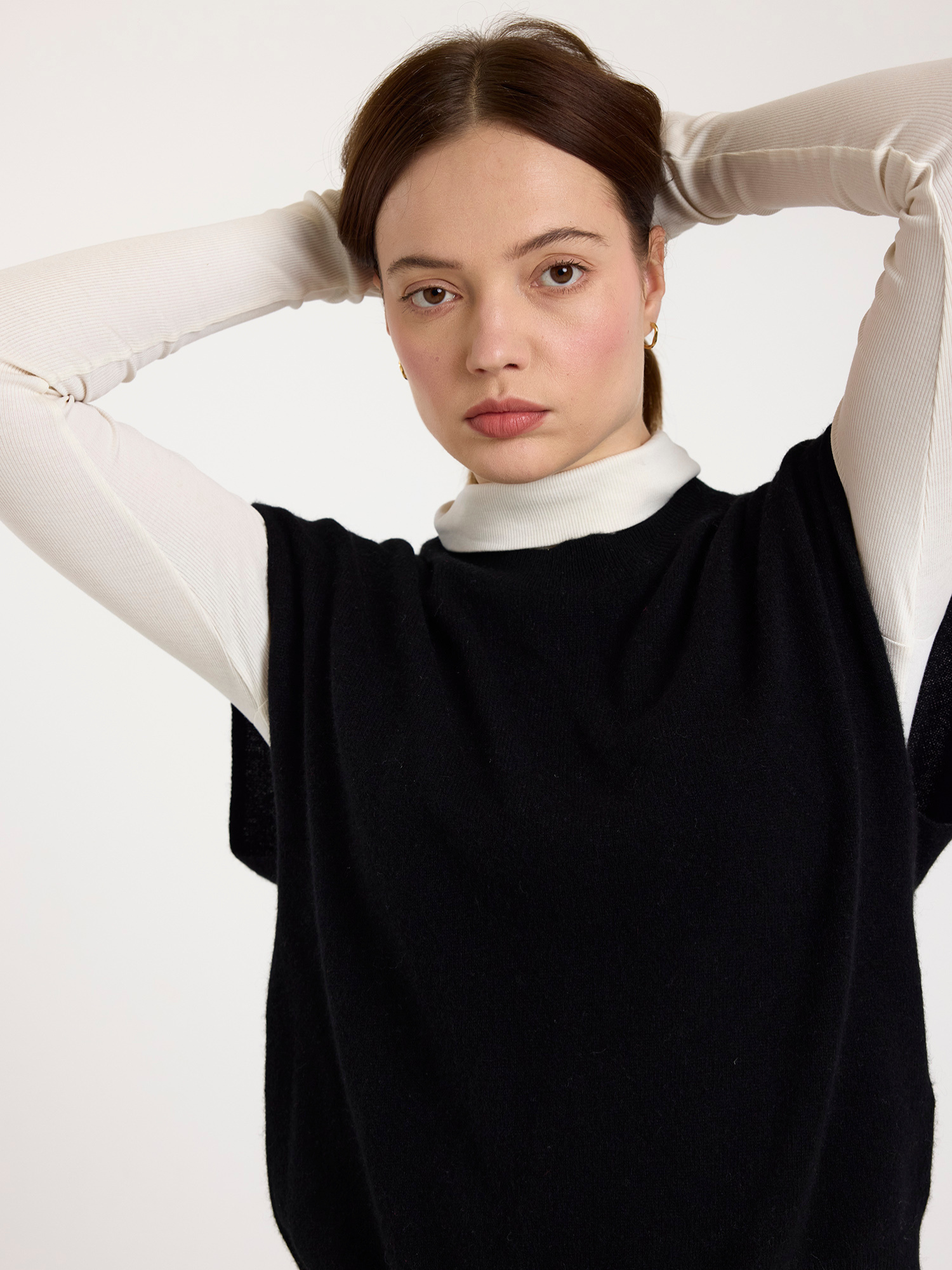 Woman wearing a black sleeveless sweater with a white polo neck underneath against a white background