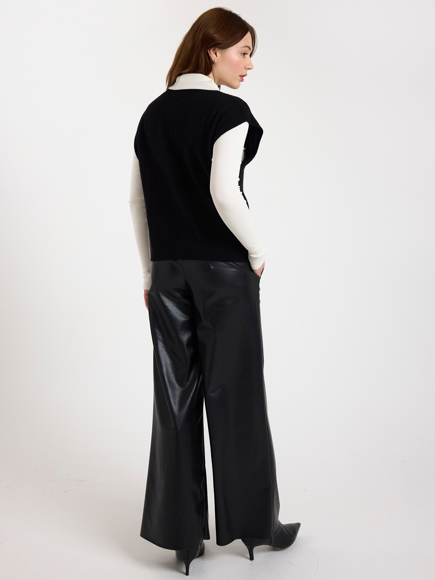 Woman wearing a black sleeveless sweater with a white polo neck underneath it with black wide-leg pants on a white background