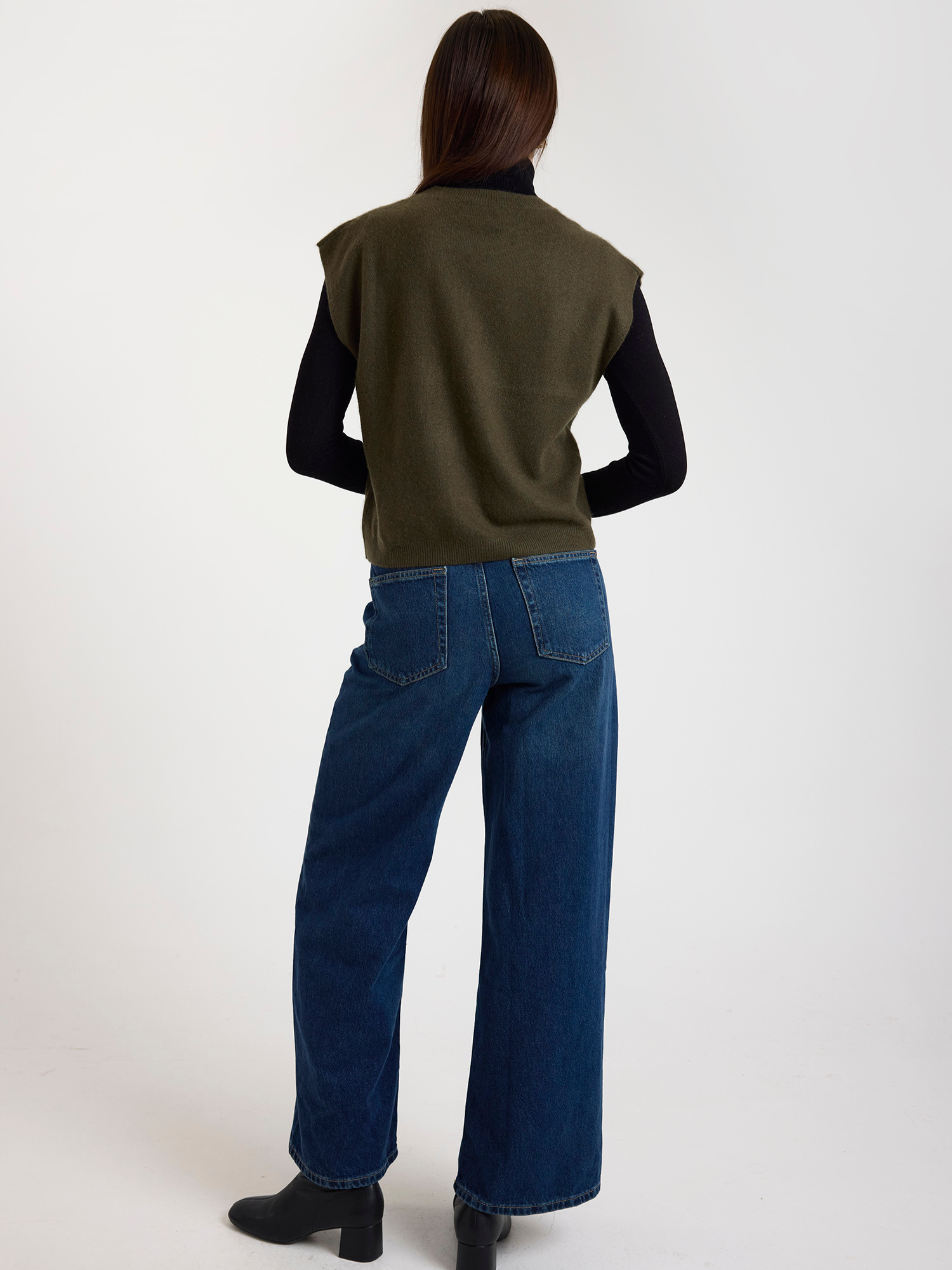 Woman wearing a green sleeveless sweater over a black turtleneck and blue jeans on a white background