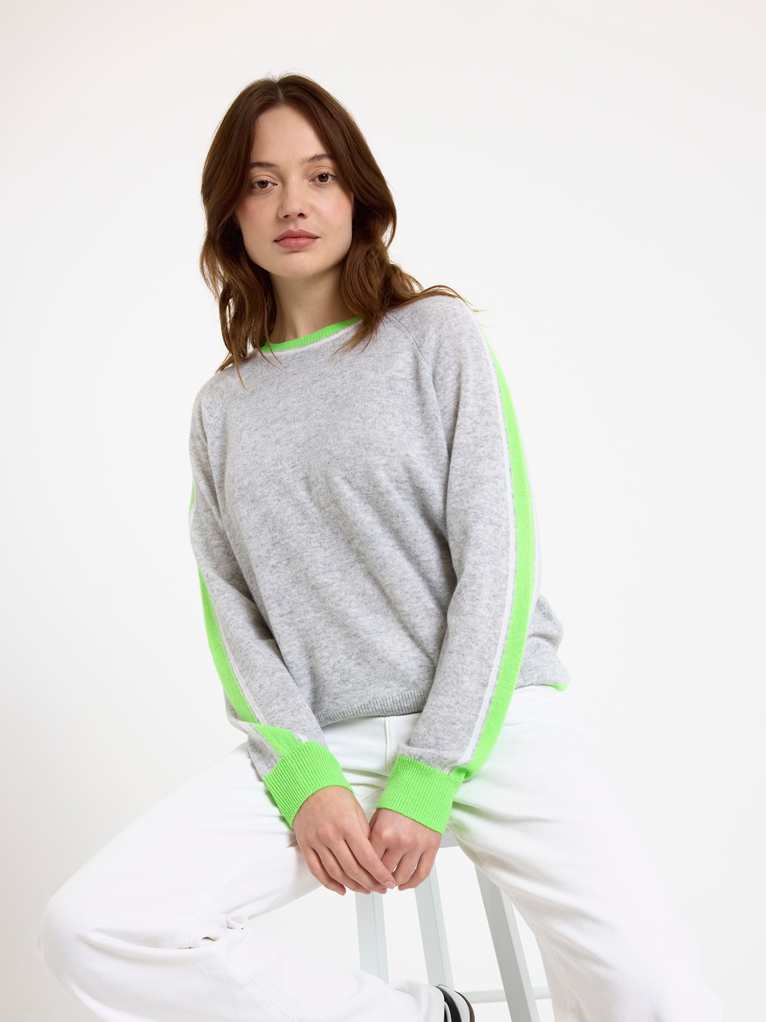 Woman wearing a grey sweater with green stripe down the sleeve and white jeans on a white background