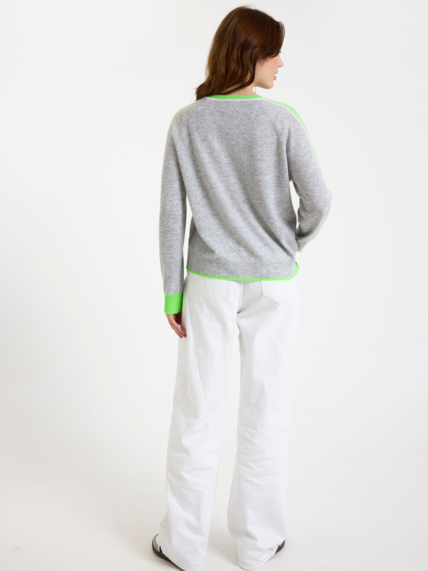 Woman wearing a grey sweater with green stripe down the sleeve and white jeans on a white background