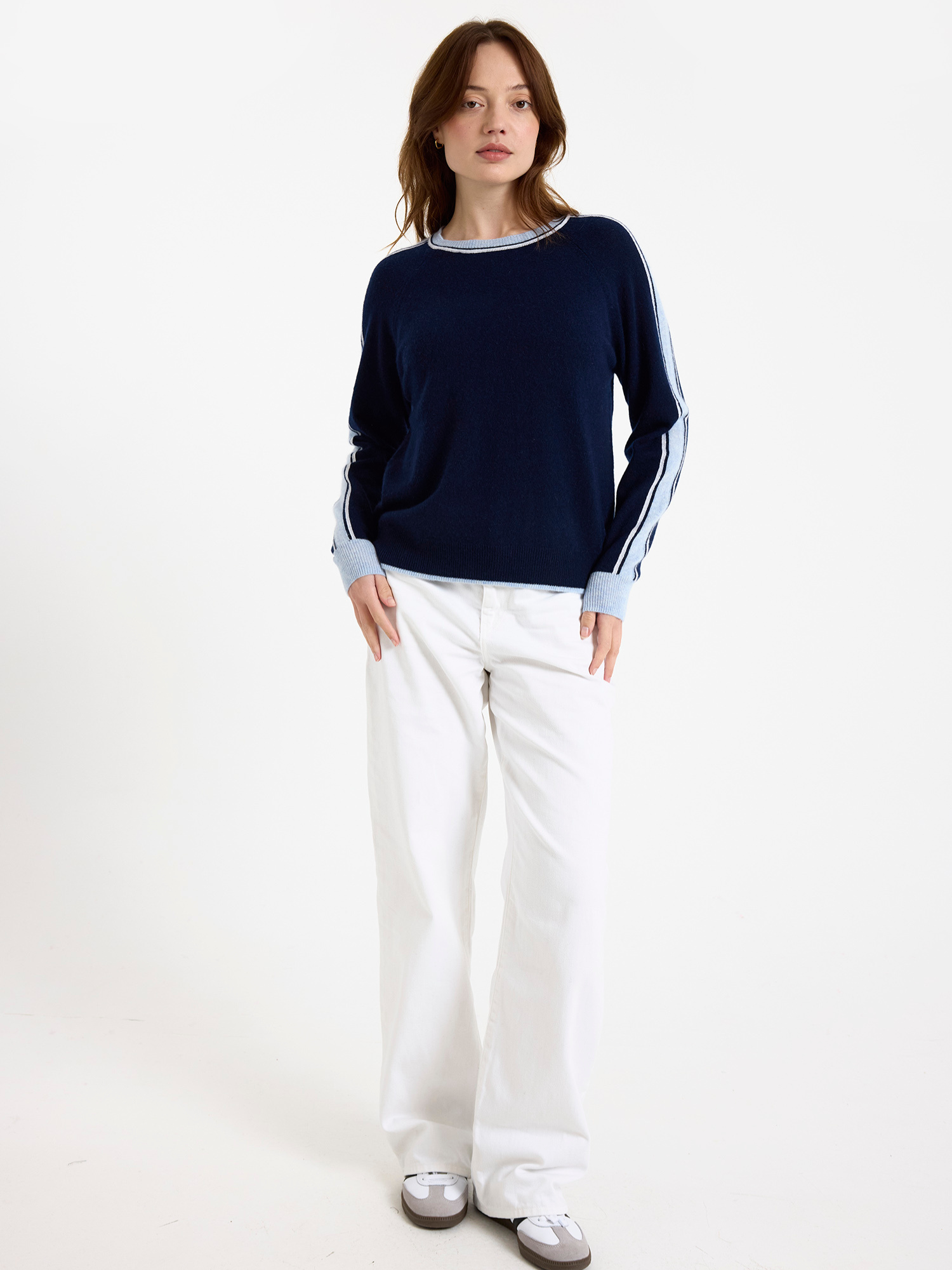 Woman wearing a navy sweater with light blue stripes down the sleeve and white jeans on a white background