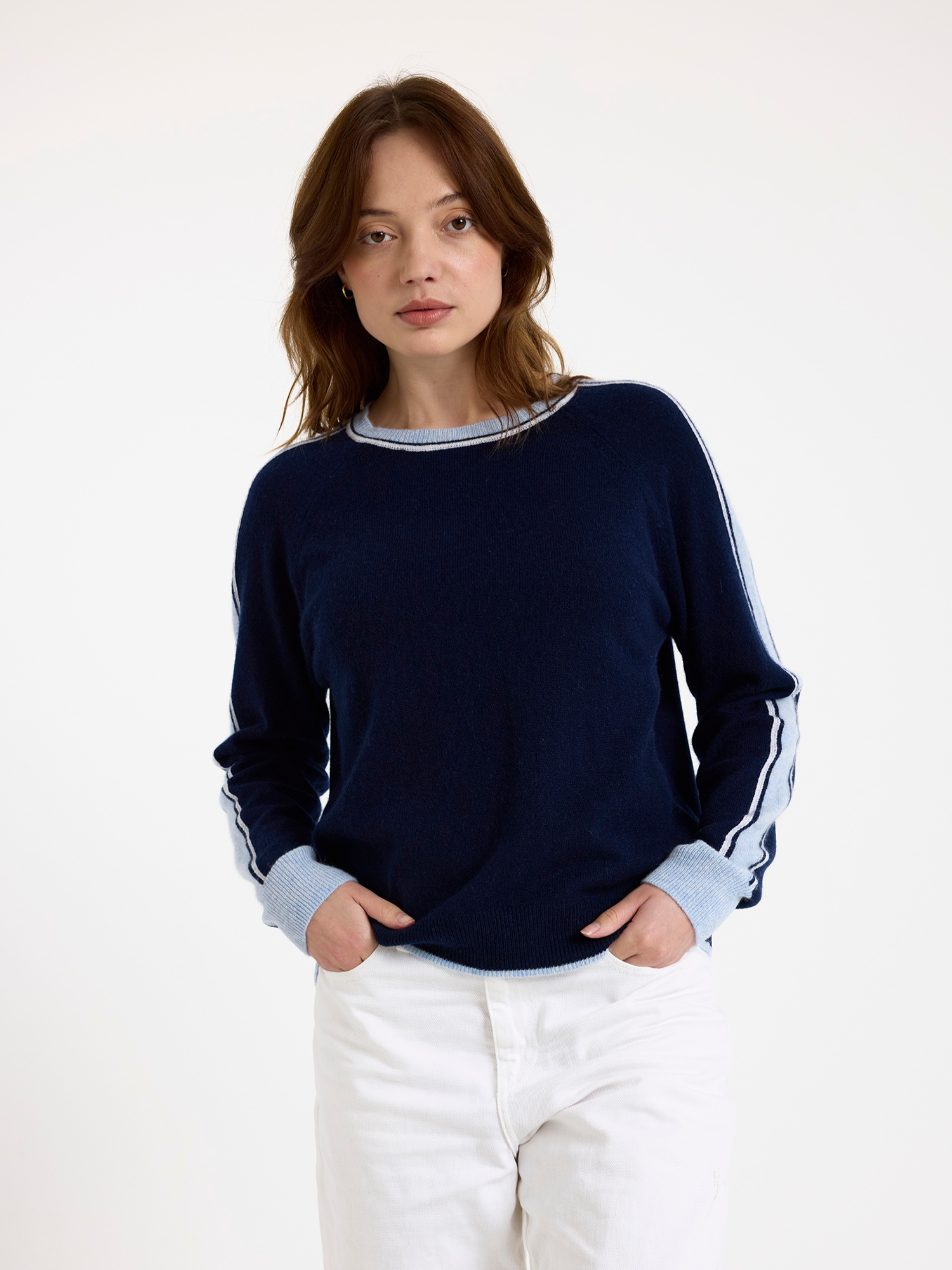 Woman wearing a navy sweater with light blue stripes down the sleeve and white jeans on a white background