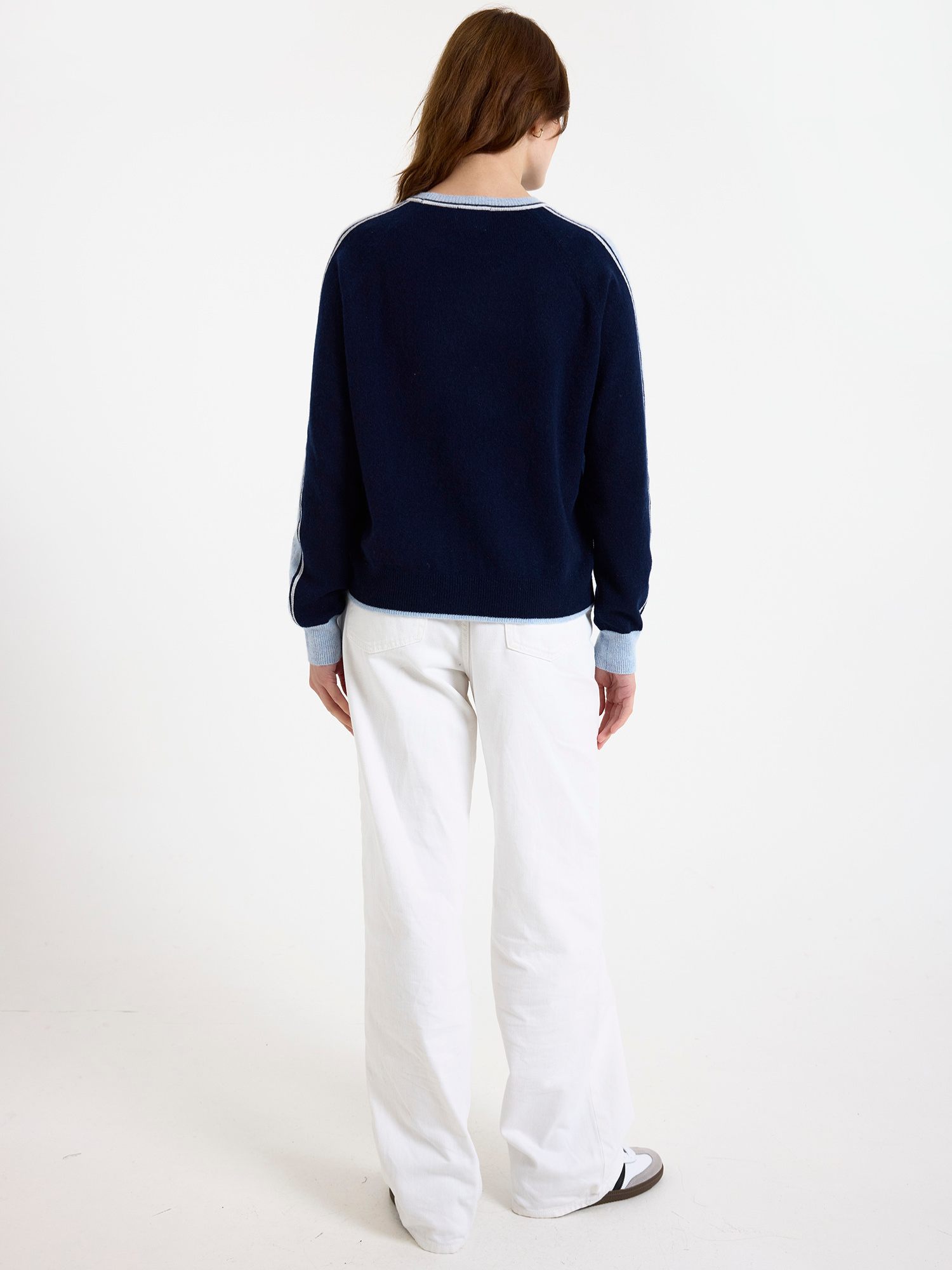 Woman wearing a navy sweater with light blue stripes down the sleeve and white jeans on a white background