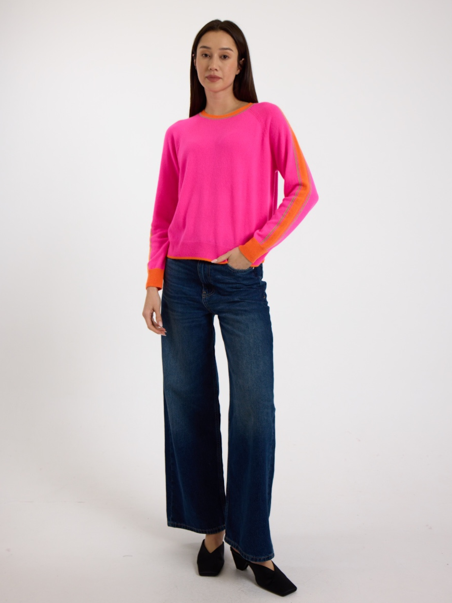 Woman wearing a bright pink sweater with orange stripes down the sleeve and blue jeans on a white background