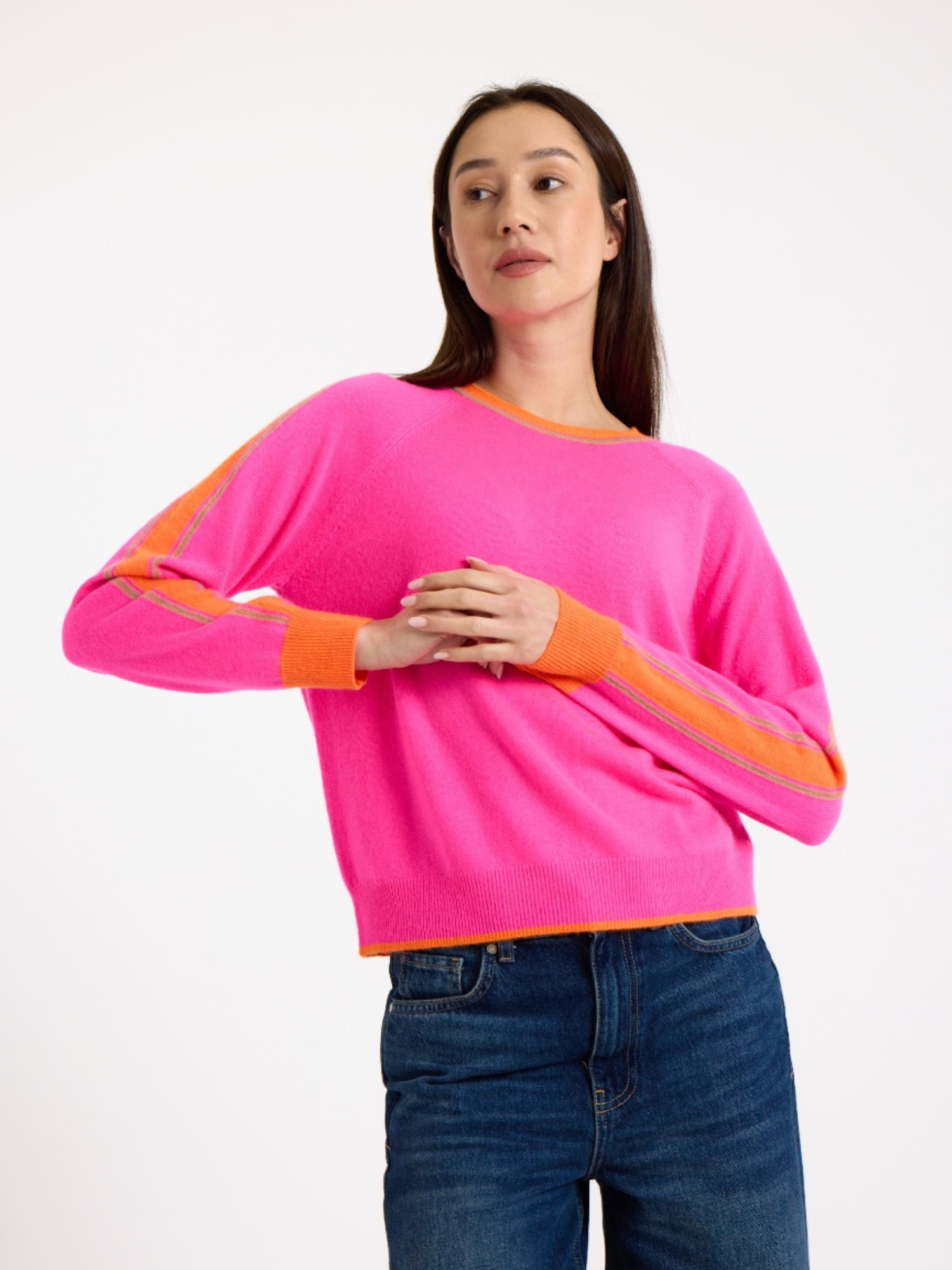 Woman wearing a bright pink sweater with orange stripes down the sleeve and blue jeans on a white background
