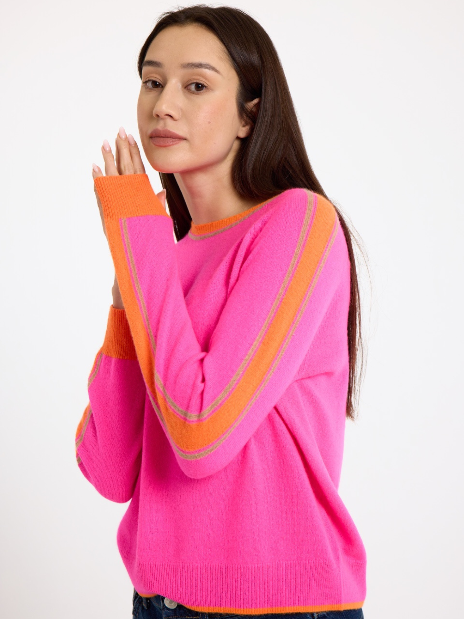 Woman wearing a bright pink sweater with orange stripes down the sleeve and blue jeans on a white background