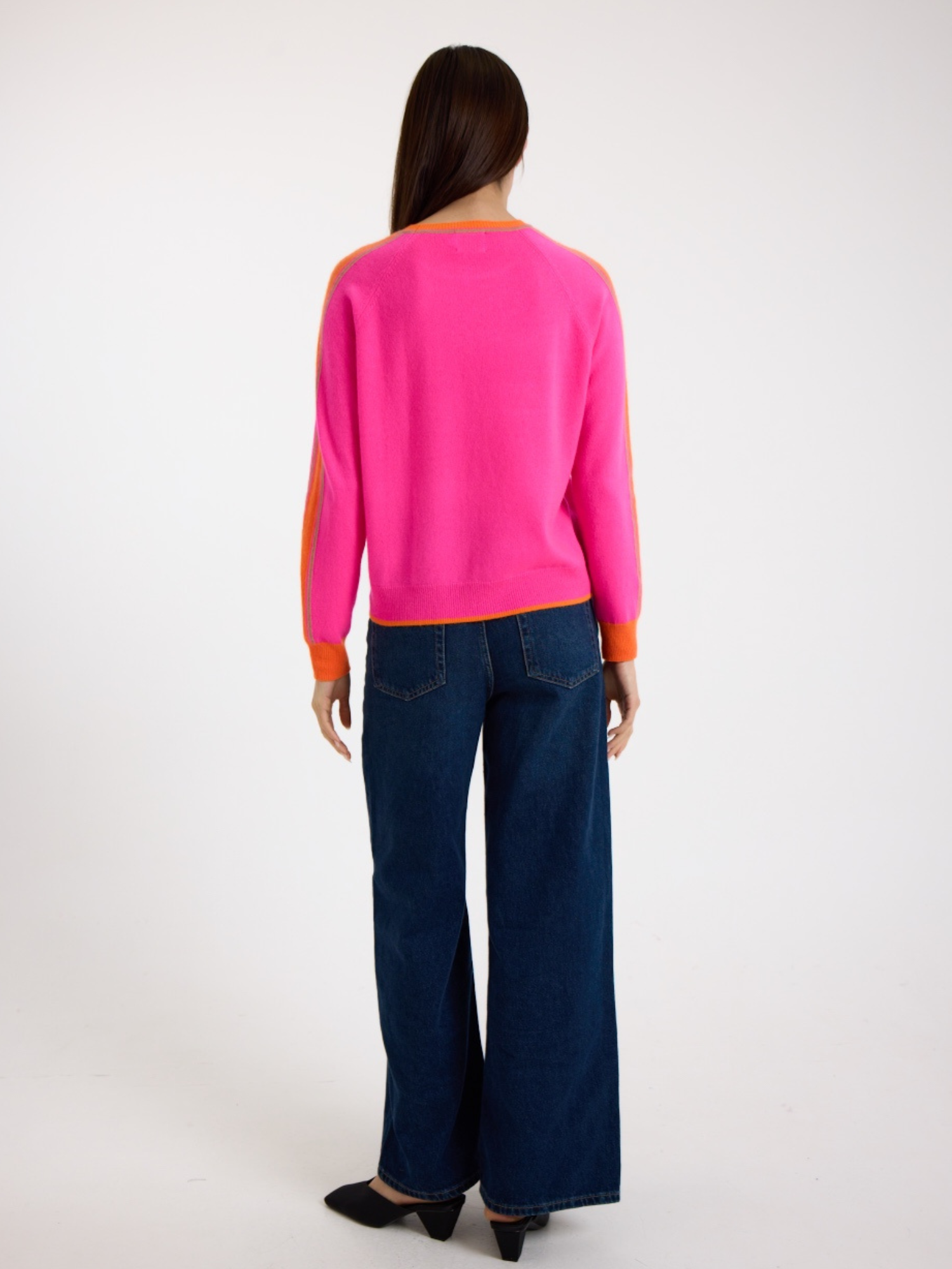 Woman wearing a bright pink sweater with orange stripes down the sleeve and blue jeans on a white background