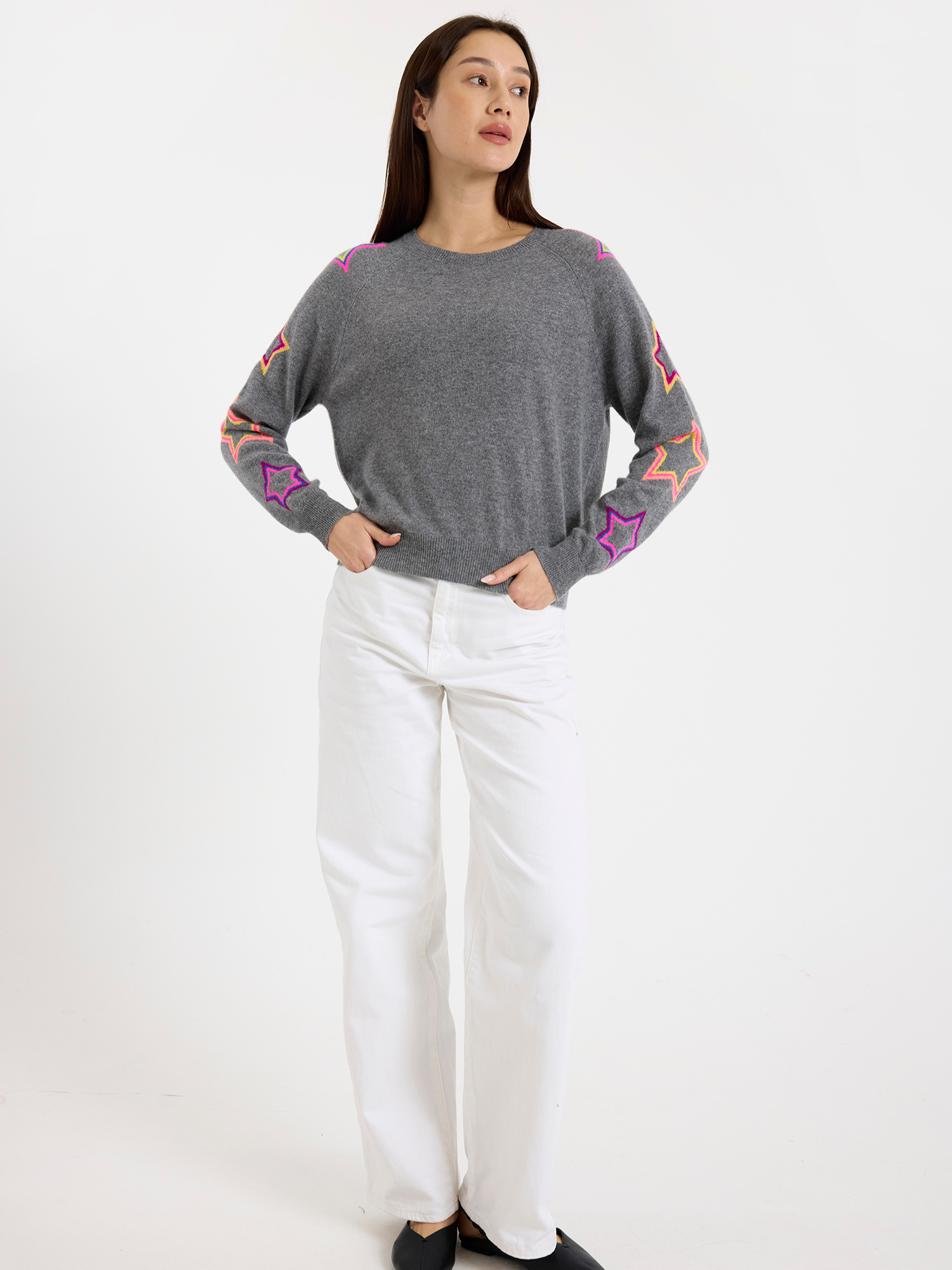 Woman wearing a grey sweater with colourful star patterns and white jeans on a white background
