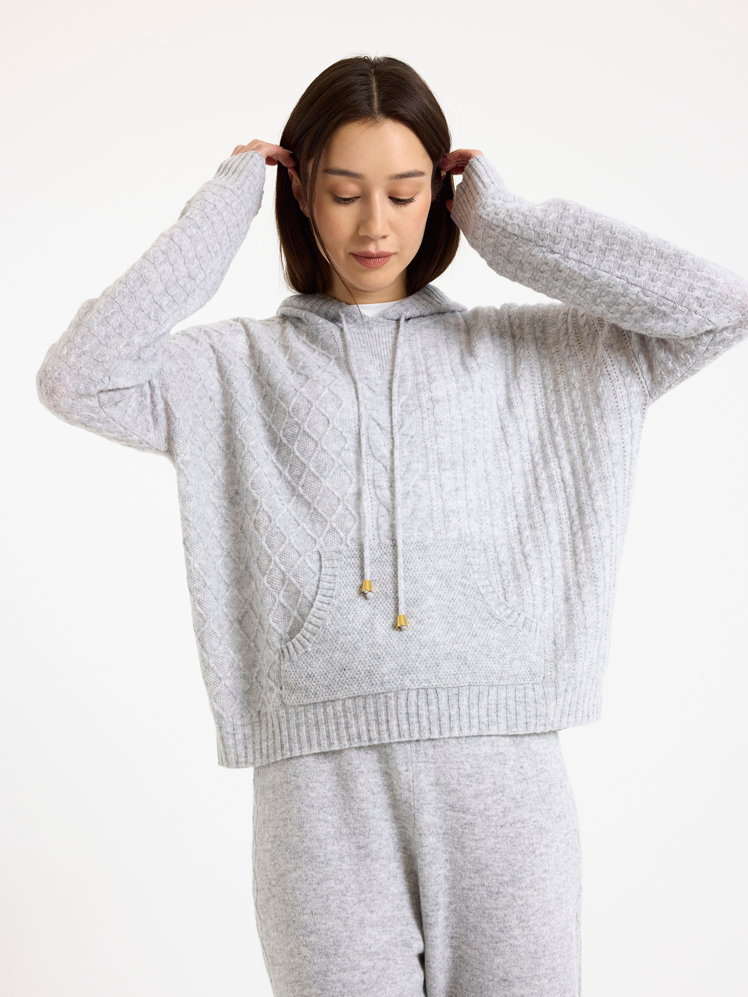 Woman wearing a grey cable knit hoodie and joggers on a white background