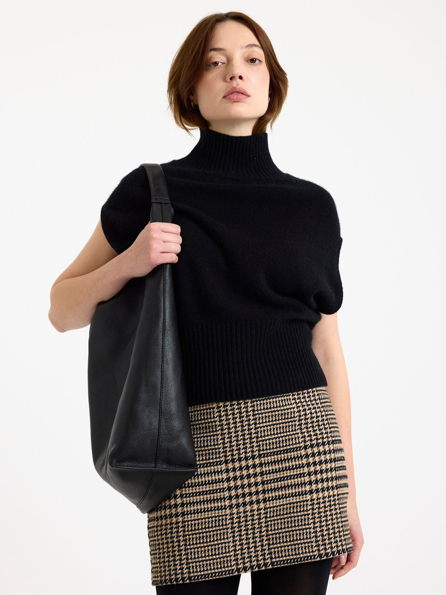 Woman wearing a black sleeveless turtleneck sweater and plaid skirt holding a black bag on a white background