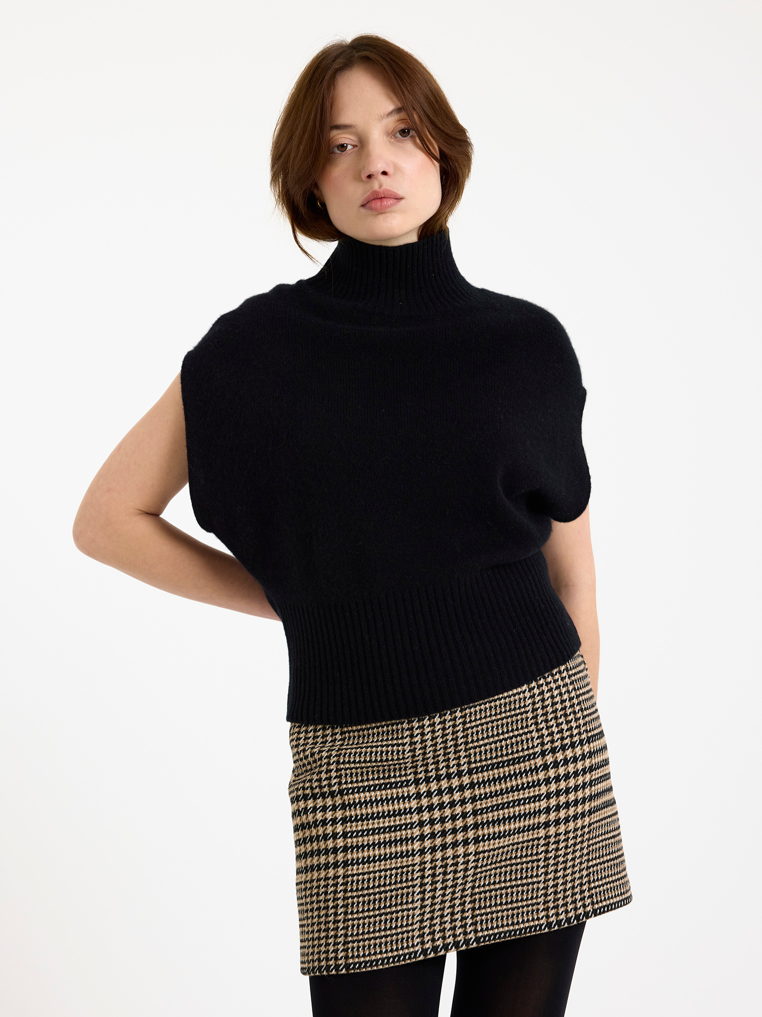 Woman wearing a black high neck sleeveless sweater and plaid skirt on a white background