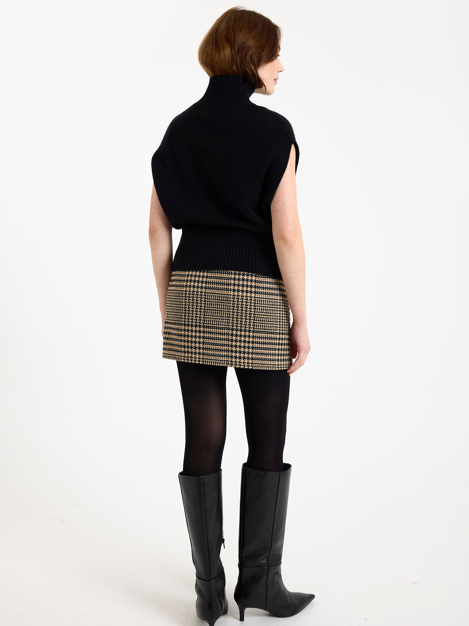 Woman wearing a black sleeveless turtleneck sweater, plaid skirt, and black boots on a white background