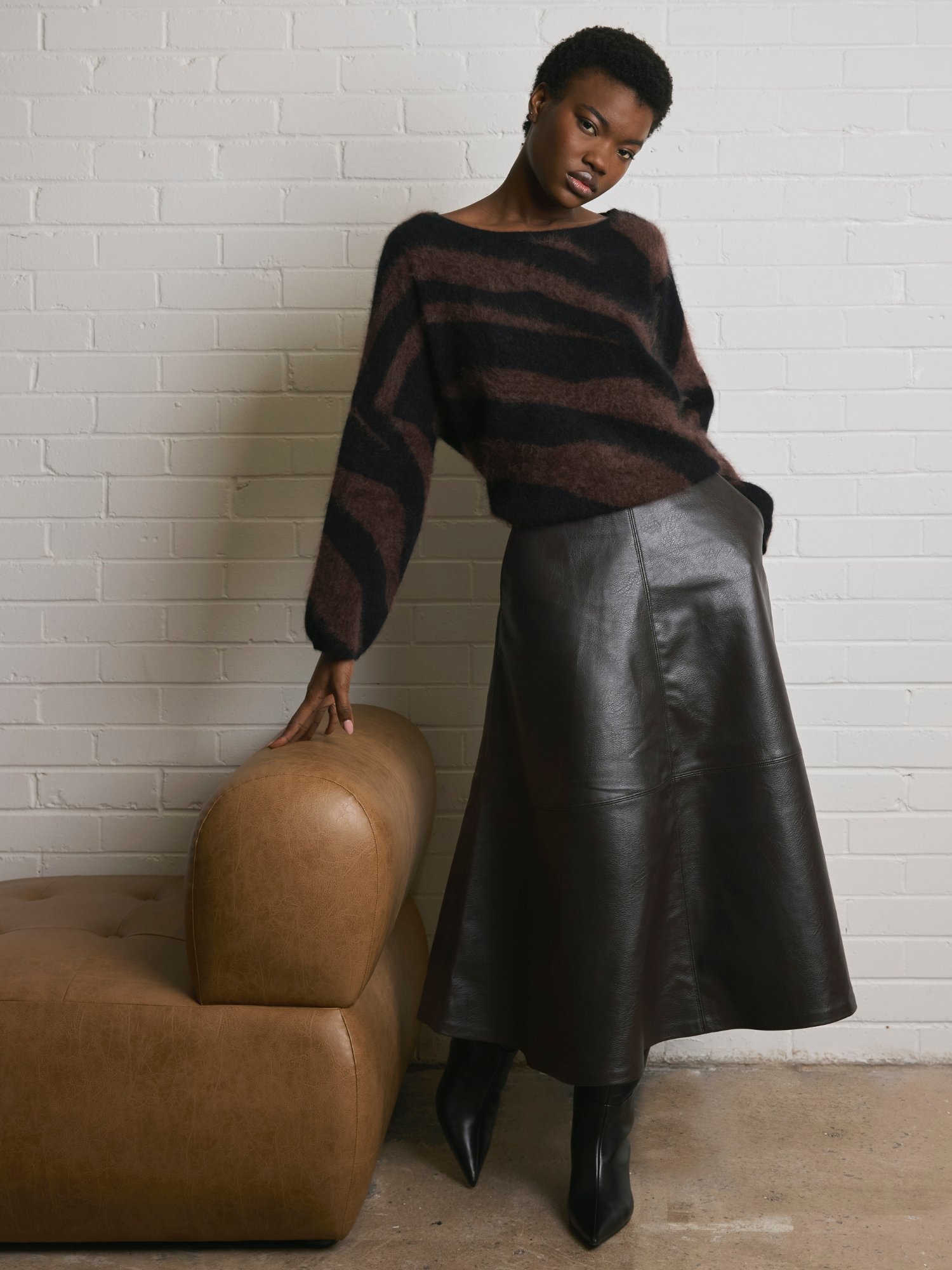 Woman in a brown and black zebra pattern sweater and brown leather skirt standing next to a brown chair against a white brick wall.