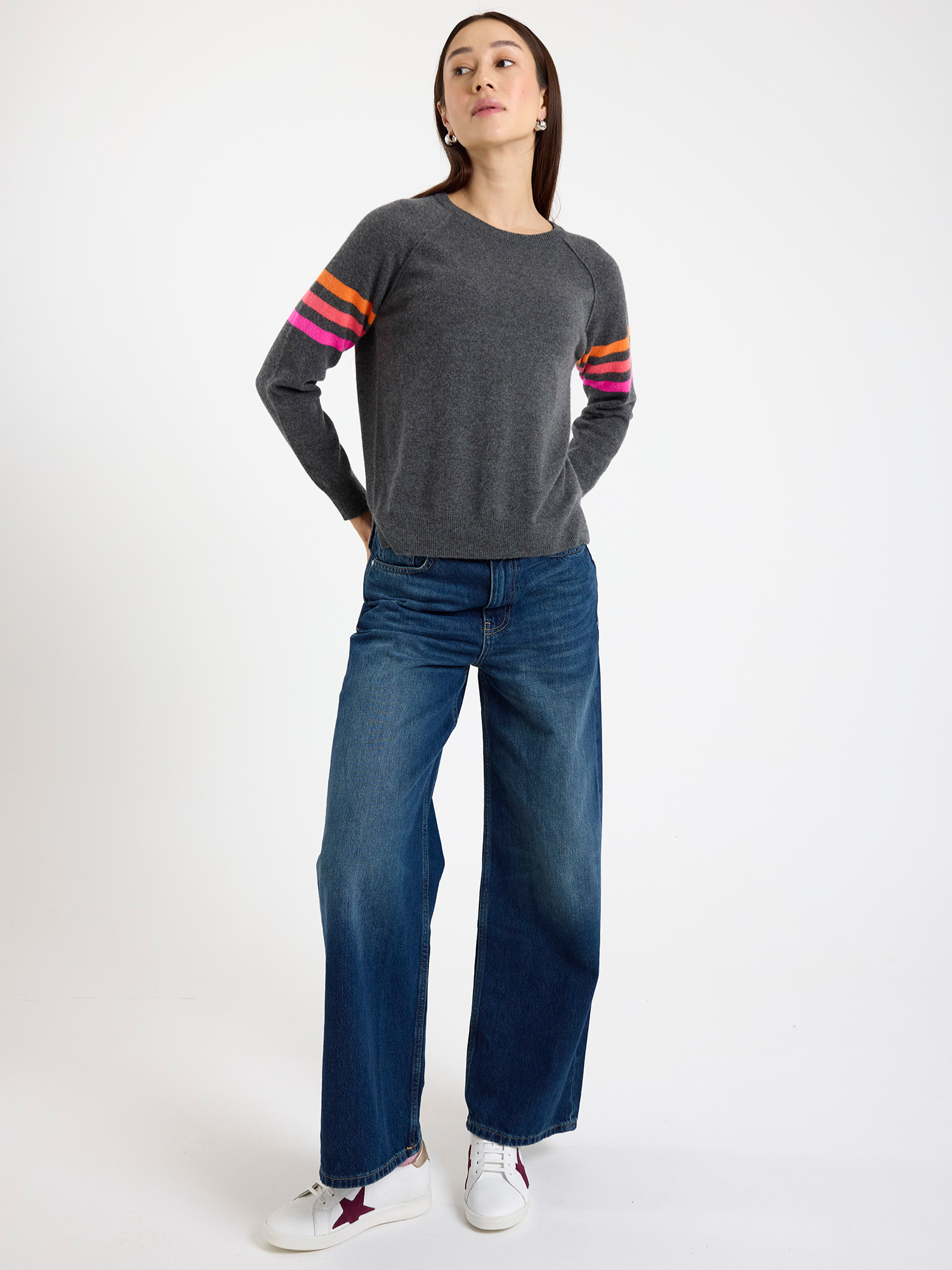 Woman wearing a dark grey sweater with colourful stripe sleeves and blue jeans on a white background