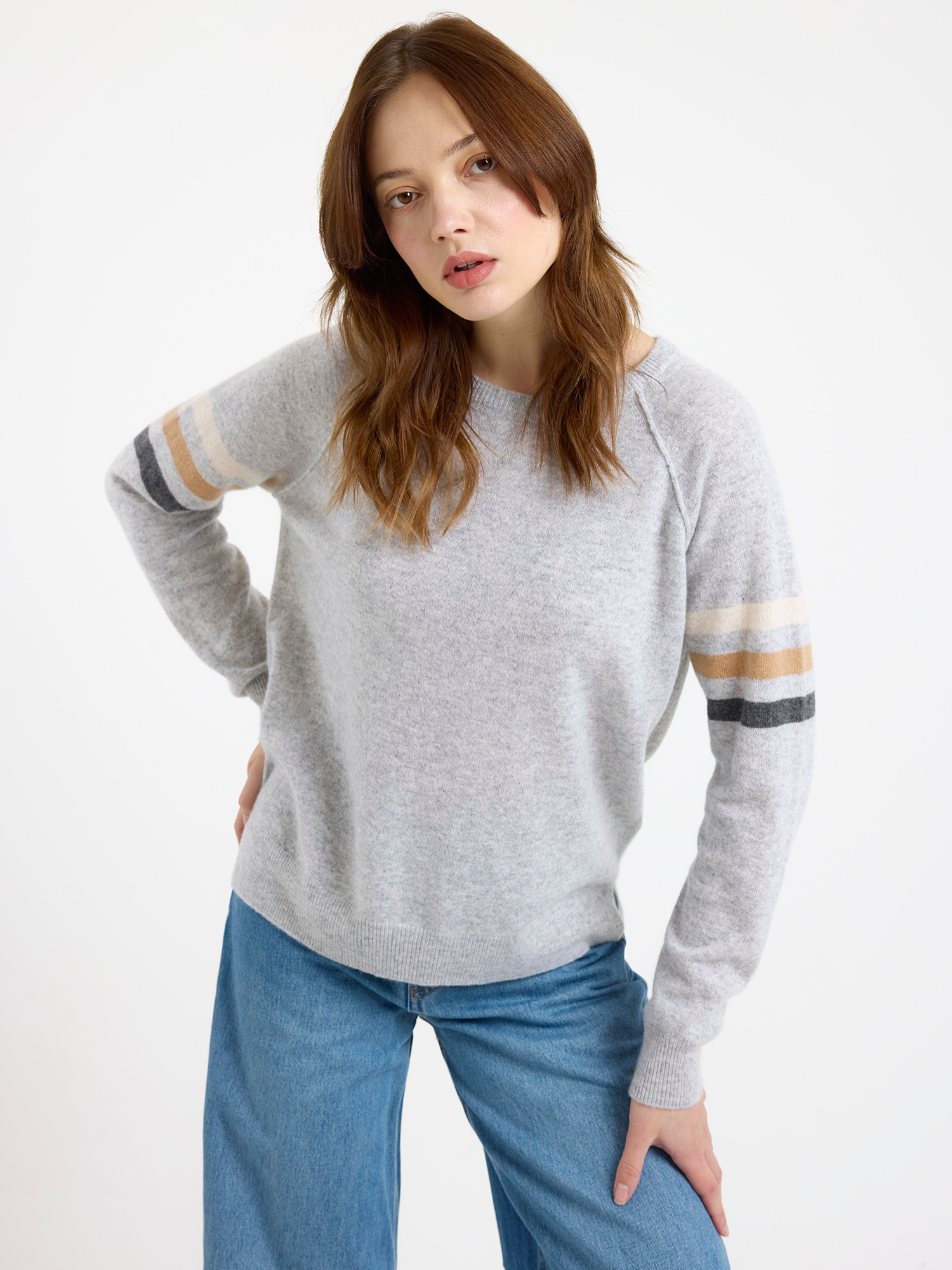 Woman wearing a grey sweater with stripe sleeves and blue jeans on a white background