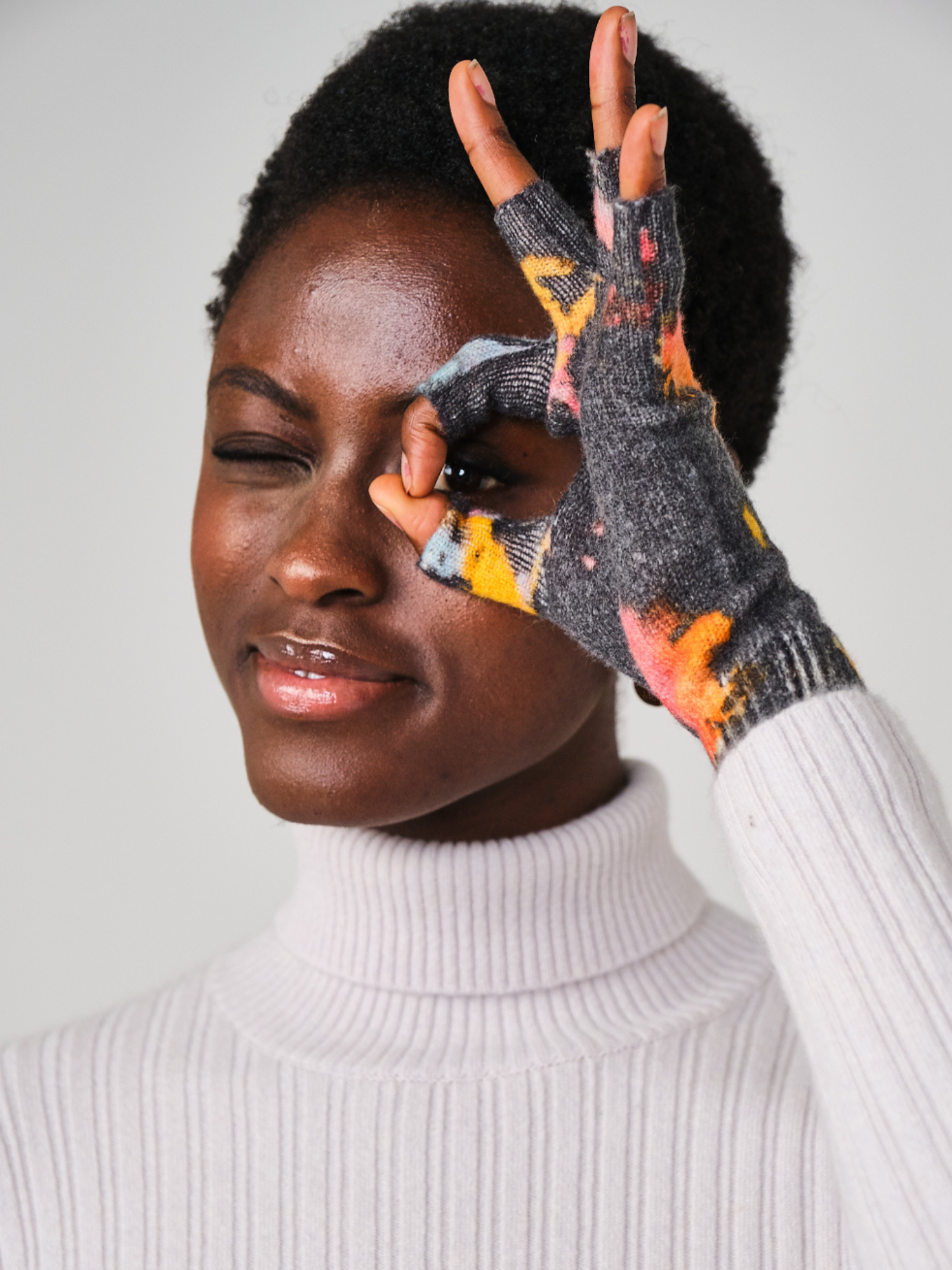Person wearing a white turtleneck and colorful gloves, holding one glove over their face.