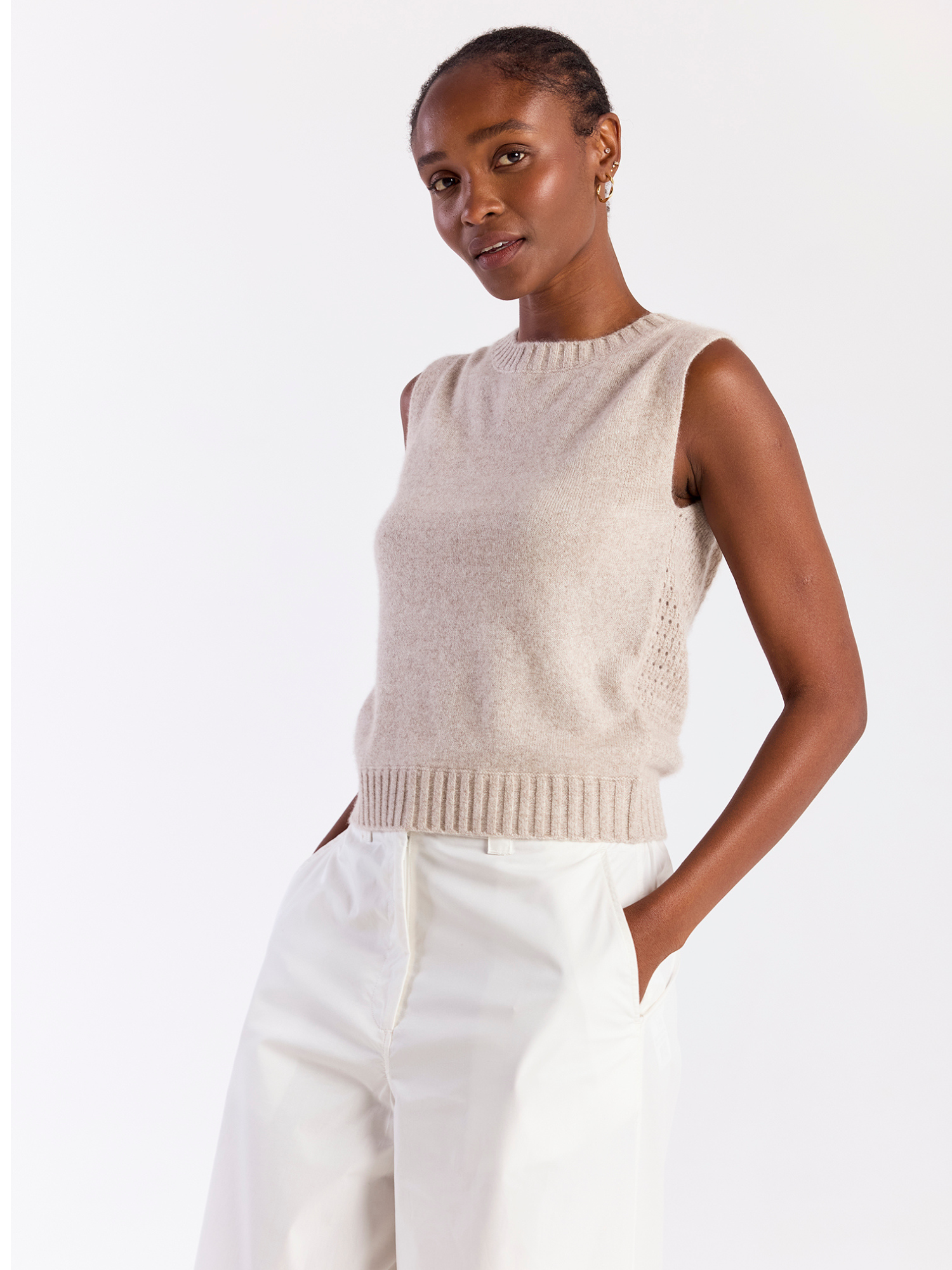Woman wearing a beige sleeveless sweater and white pants on a white background