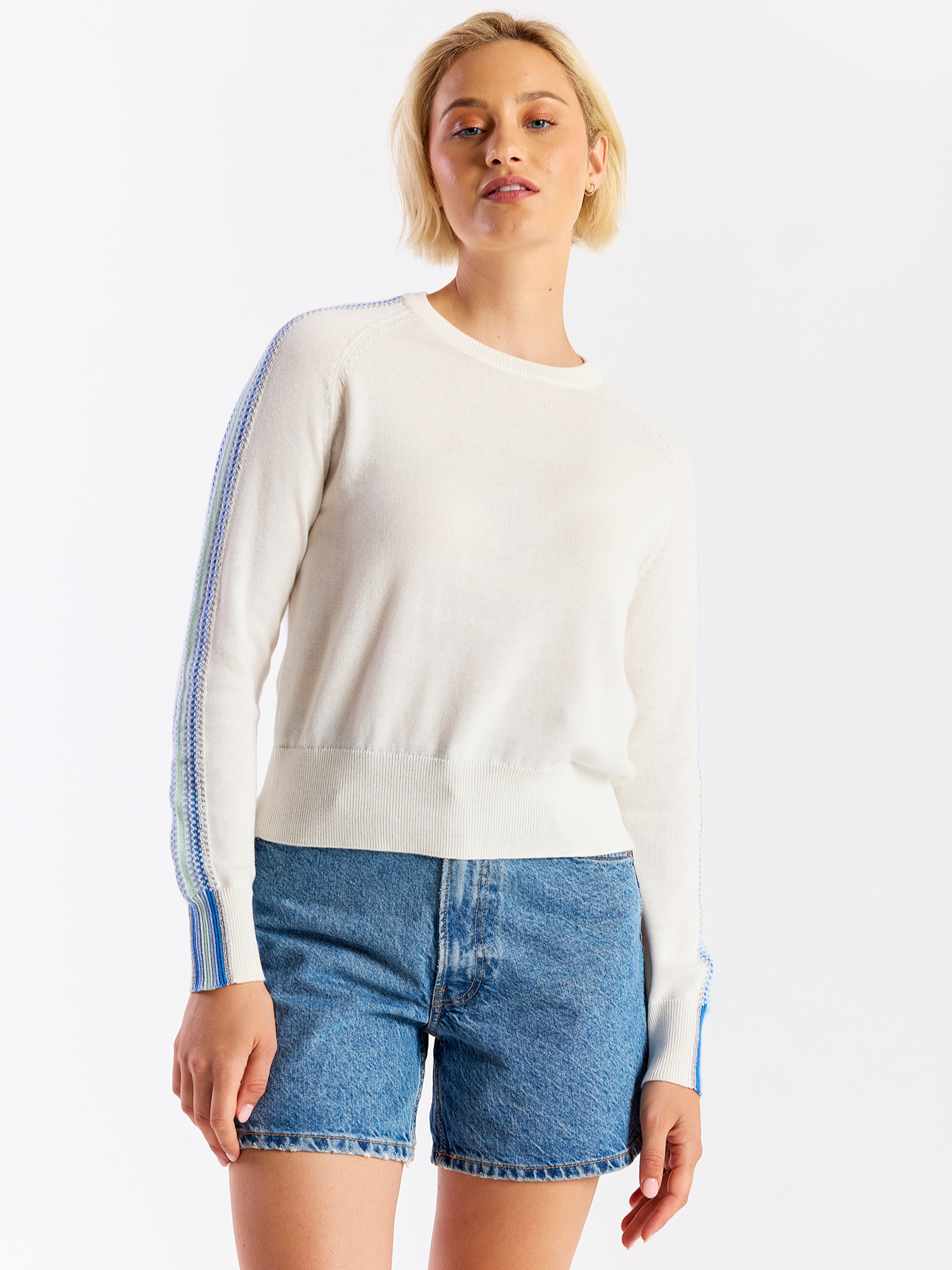 Woman wearing a white sweater with blue accents and denim shorts on a white background