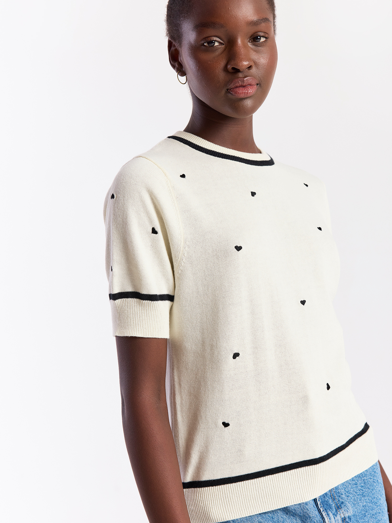 Woman wearing a cream sweater with black heart patterns and black trim on a white background