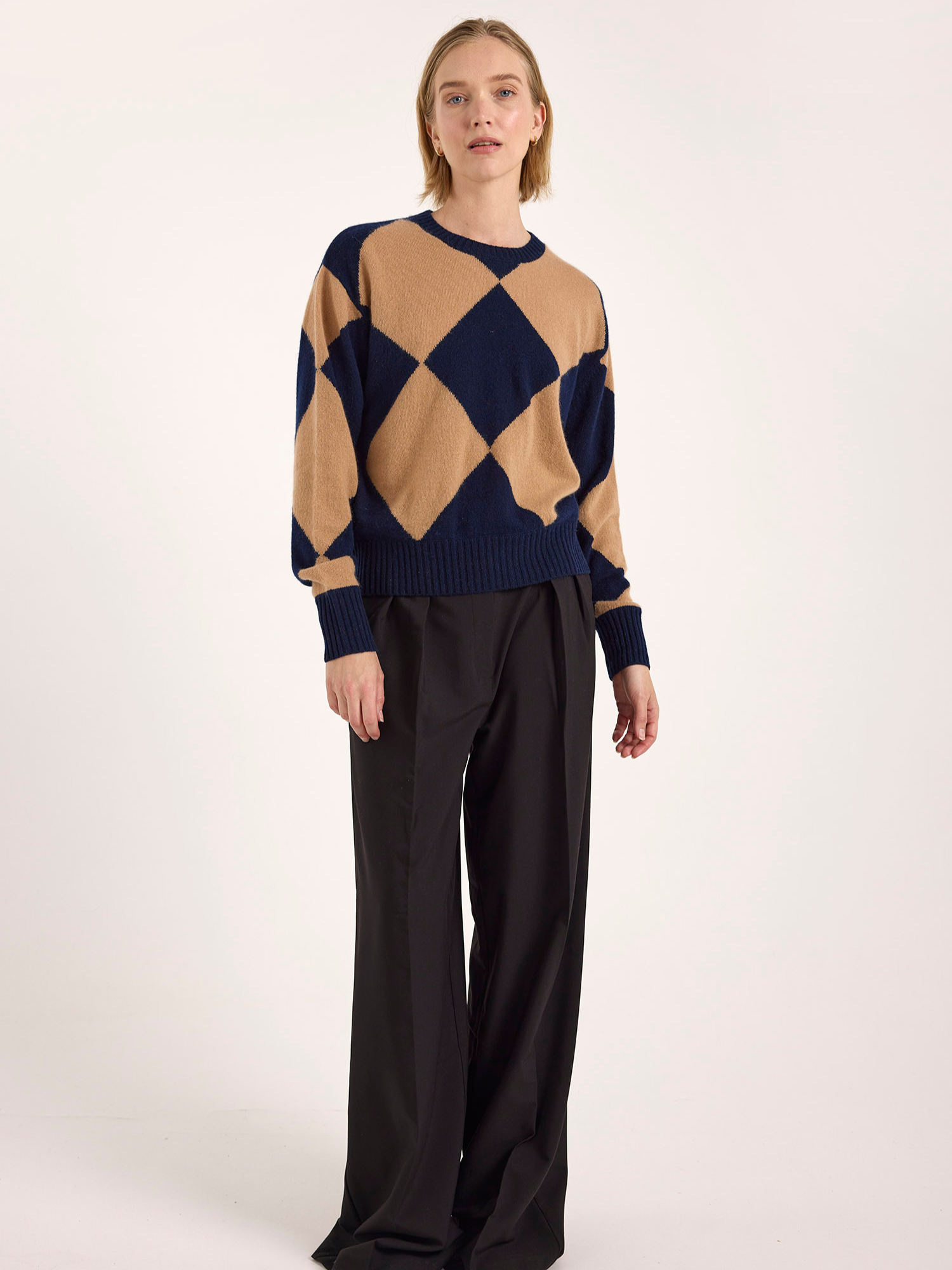 Person wearing a navy and beige argyle sweater with black pants on a white background