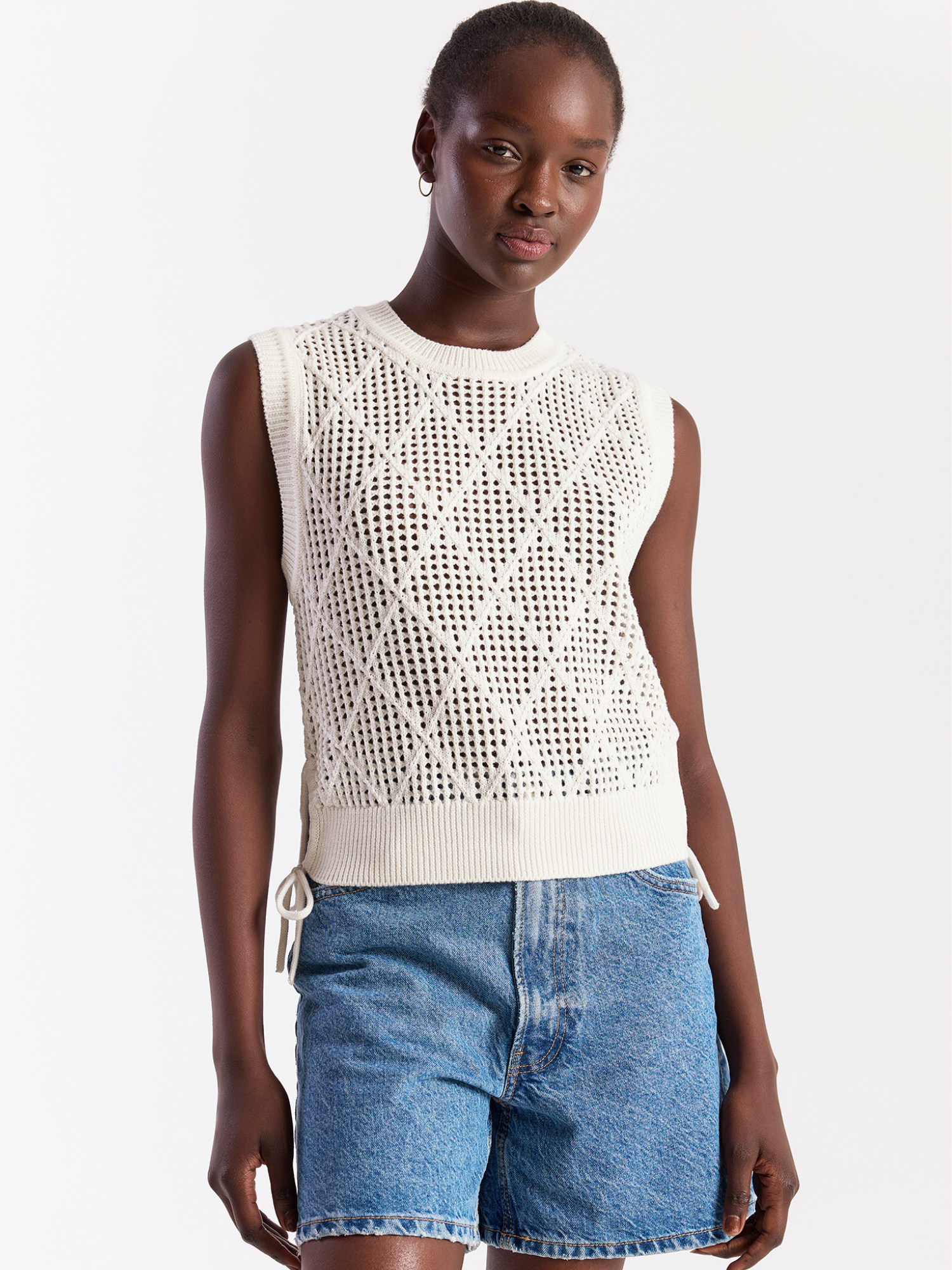 Woman wearing a white sleeveless crochet top and blue shorts on a white background